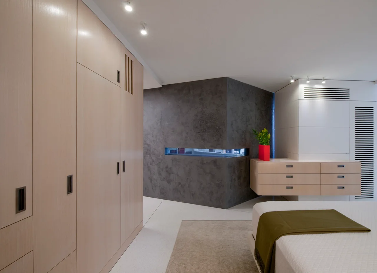 A modern bedroom features custom light wood wardrobes, a floating dresser, and a textured charcoal accent wall with a narrow horizontal window.