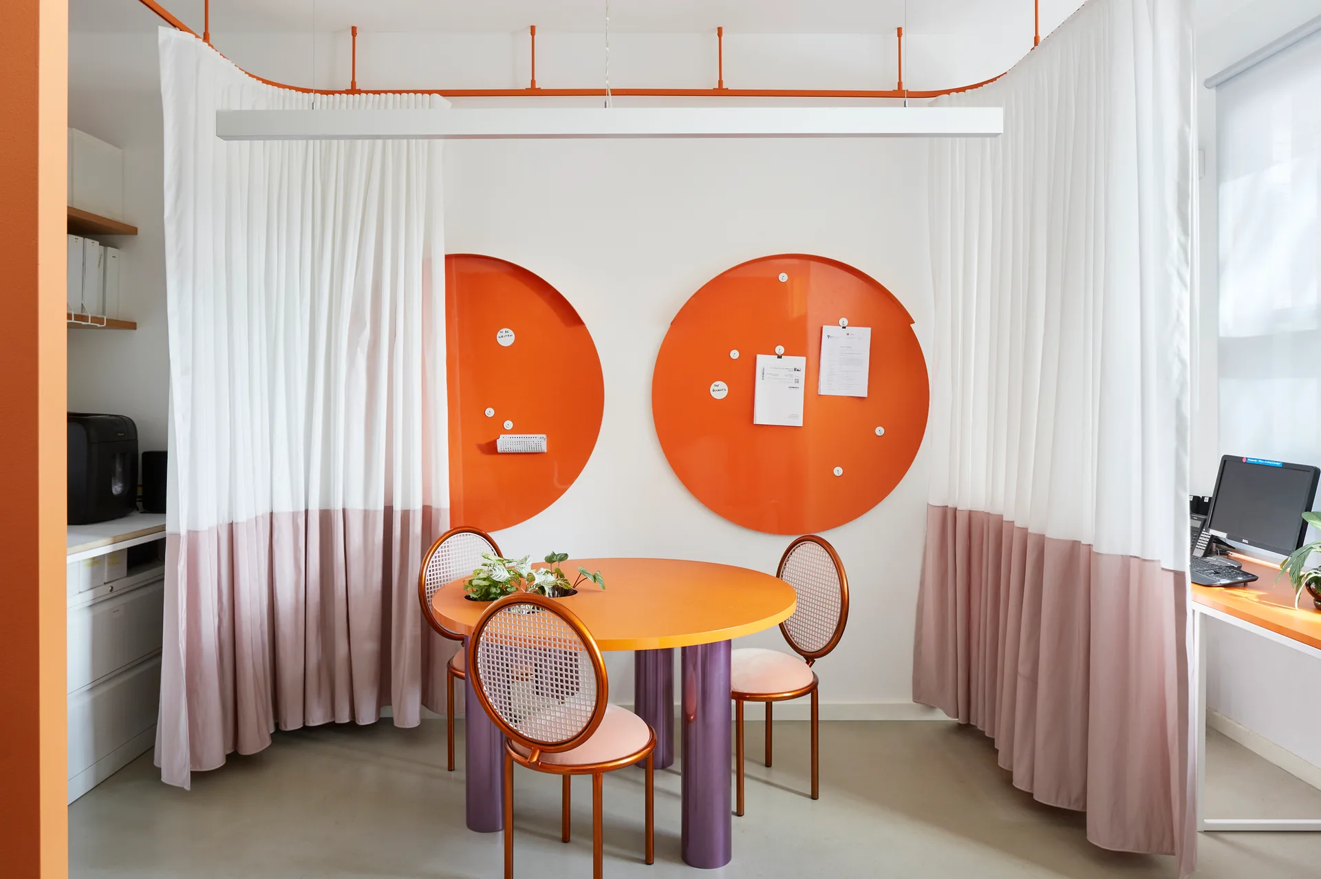 An office workspace features a round orange table surrounded by mesh-back chairs and flanked by white and mauve partition curtains hung on ceiling tracks.