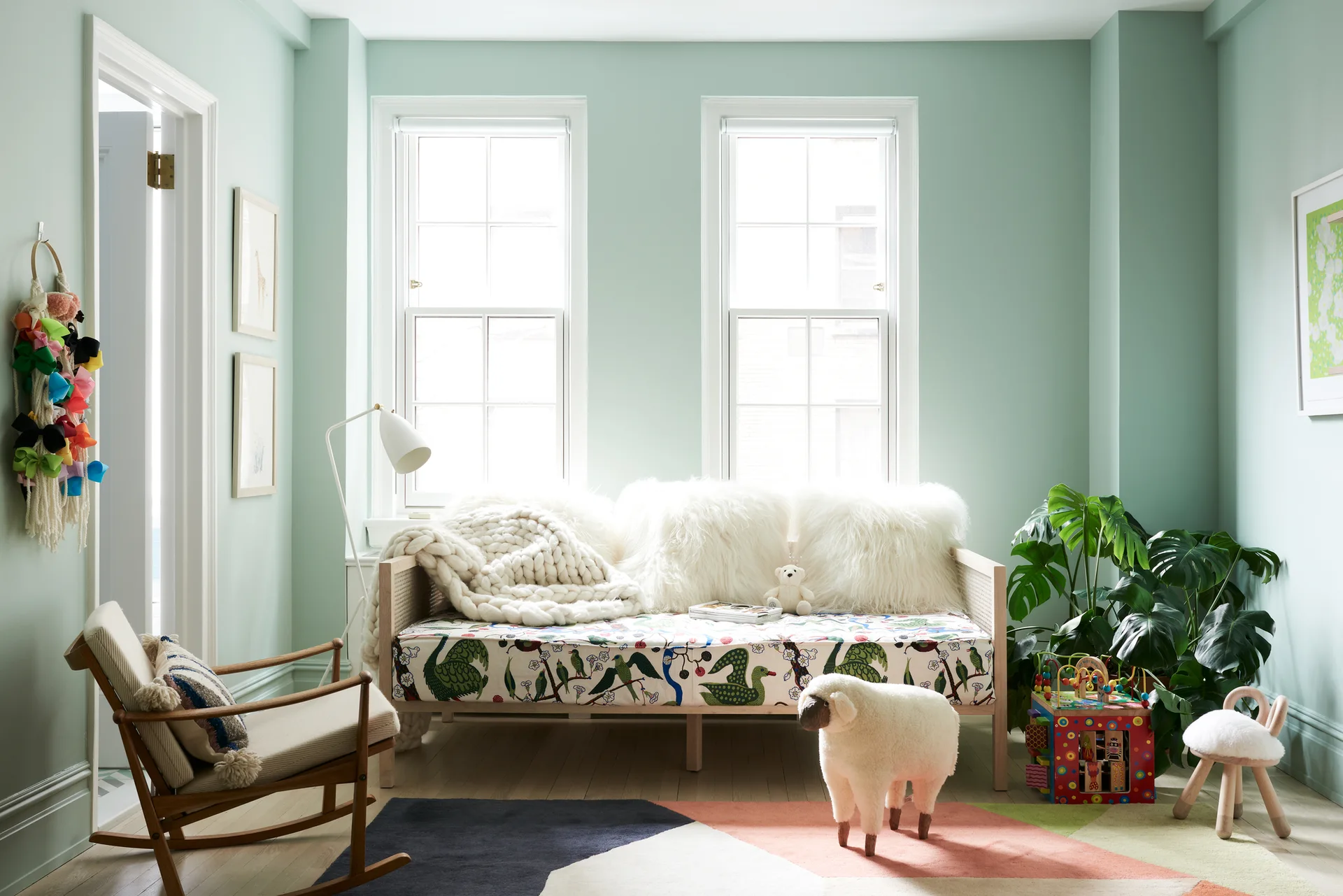 A mint green kids playroom features a rattan daybed with botanical cushions, fluffy pillows, and a sheep-shaped stool.