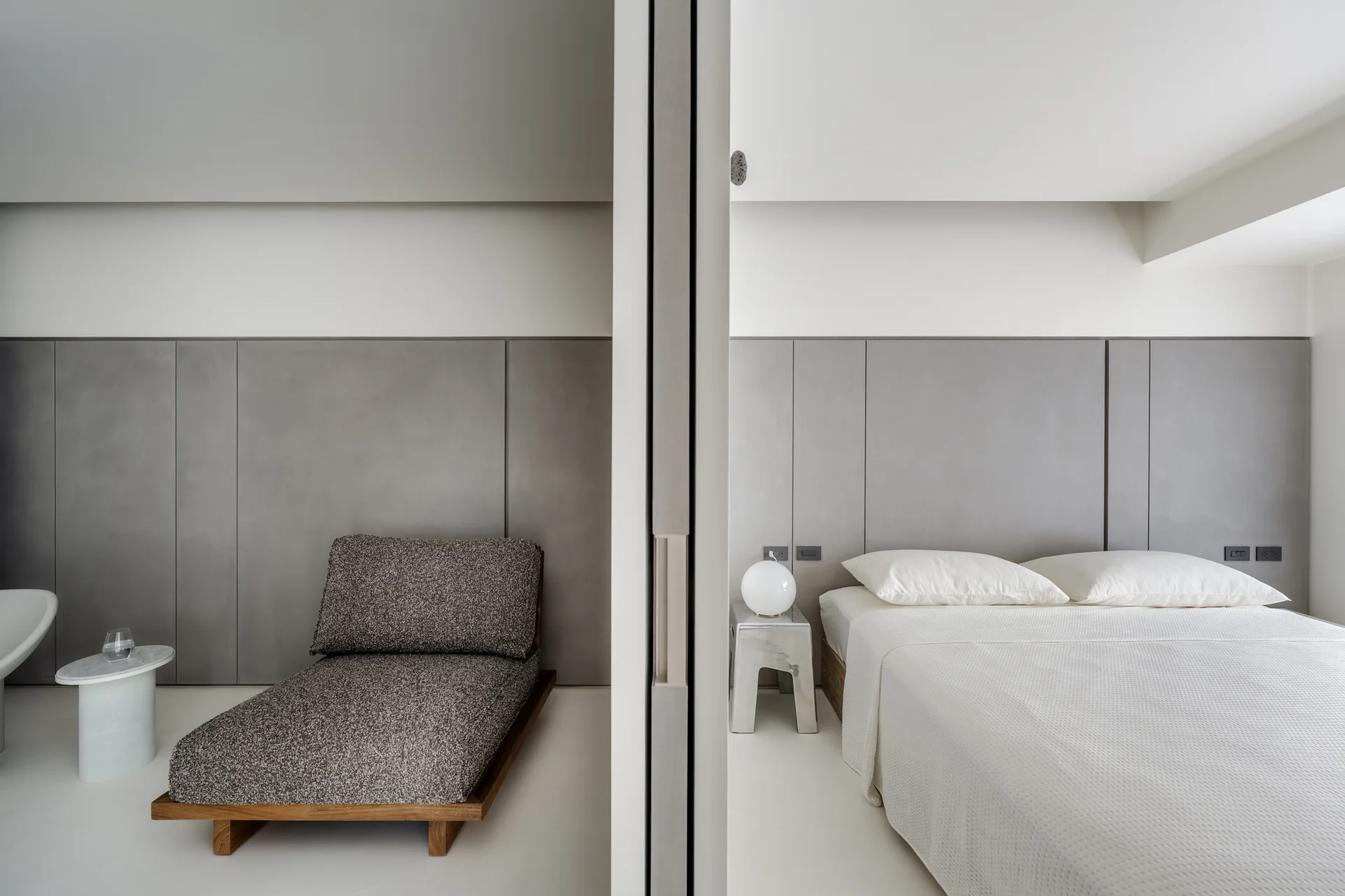 A minimalist bedroom and lounge space divided by a white partition wall, featuring grey wall paneling, a low bed, and a textured chaise lounge.