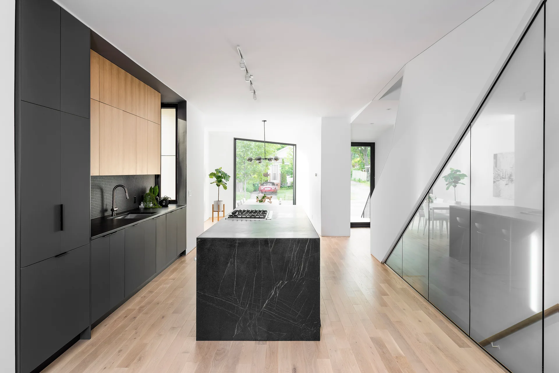 A contemporary kitchen features a dark veined stone island, matte black and wood cabinetry, light wood floors, and a staircase with a glass railing.
