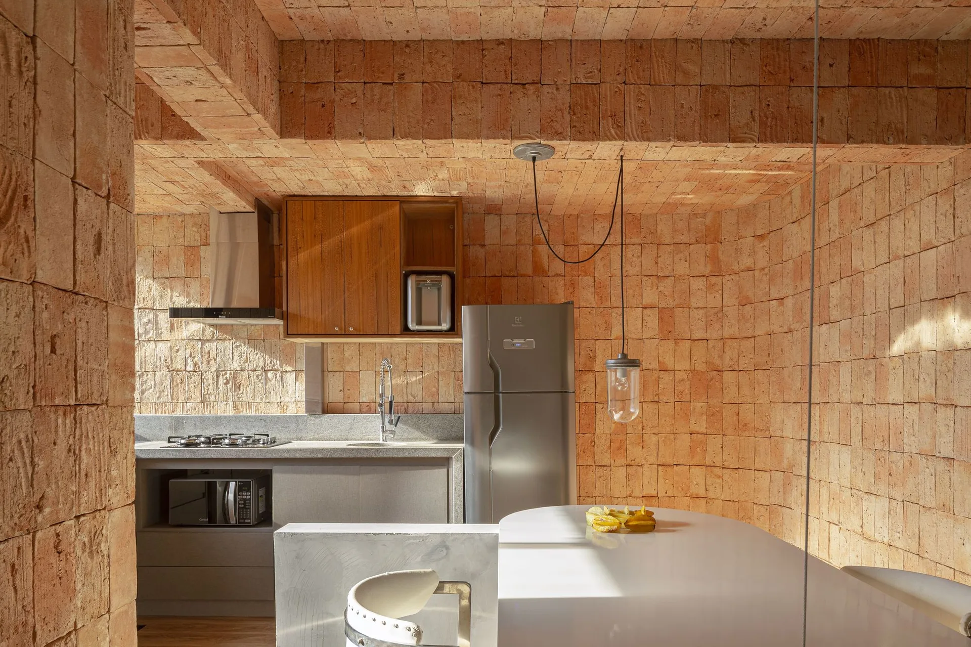 A modern kitchen features exposed terracotta brick walls and ceiling, gray cabinets, stainless steel appliances, and a white dining table.