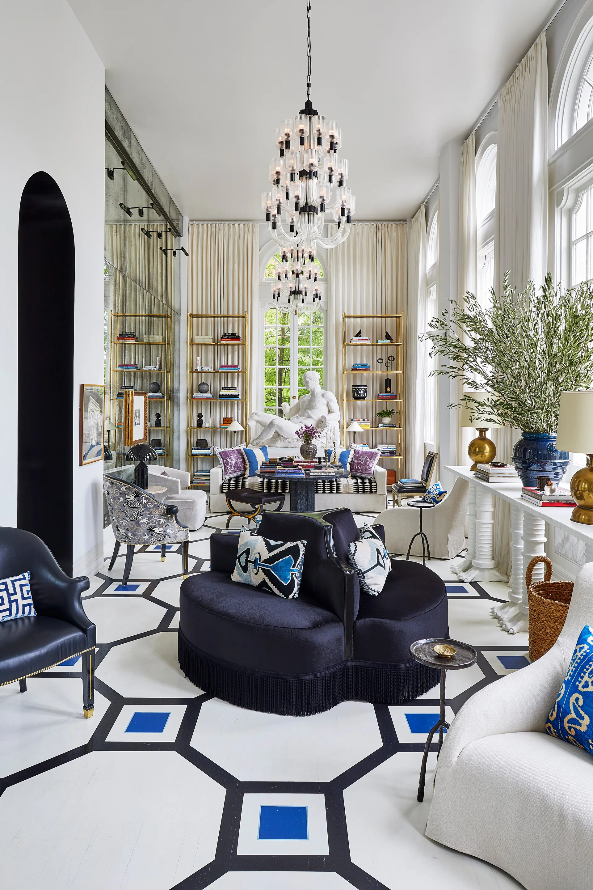 A spacious living room features a black and white geometric patterned floor, a central circular black velvet sofa, and tall arched windows.