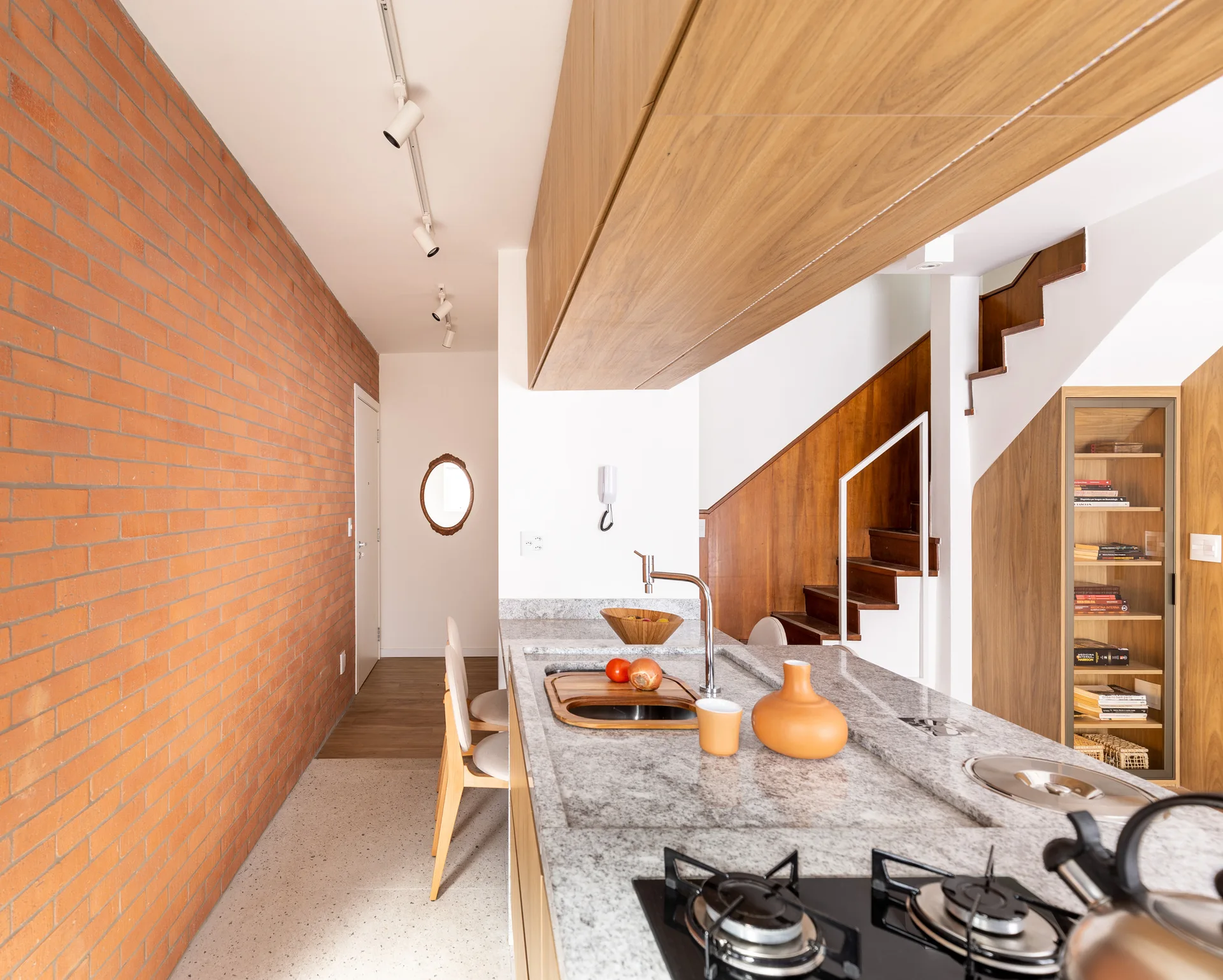 A modern kitchen features a long granite island, an exposed red brick wall, wood cabinetry, and a wooden staircase.