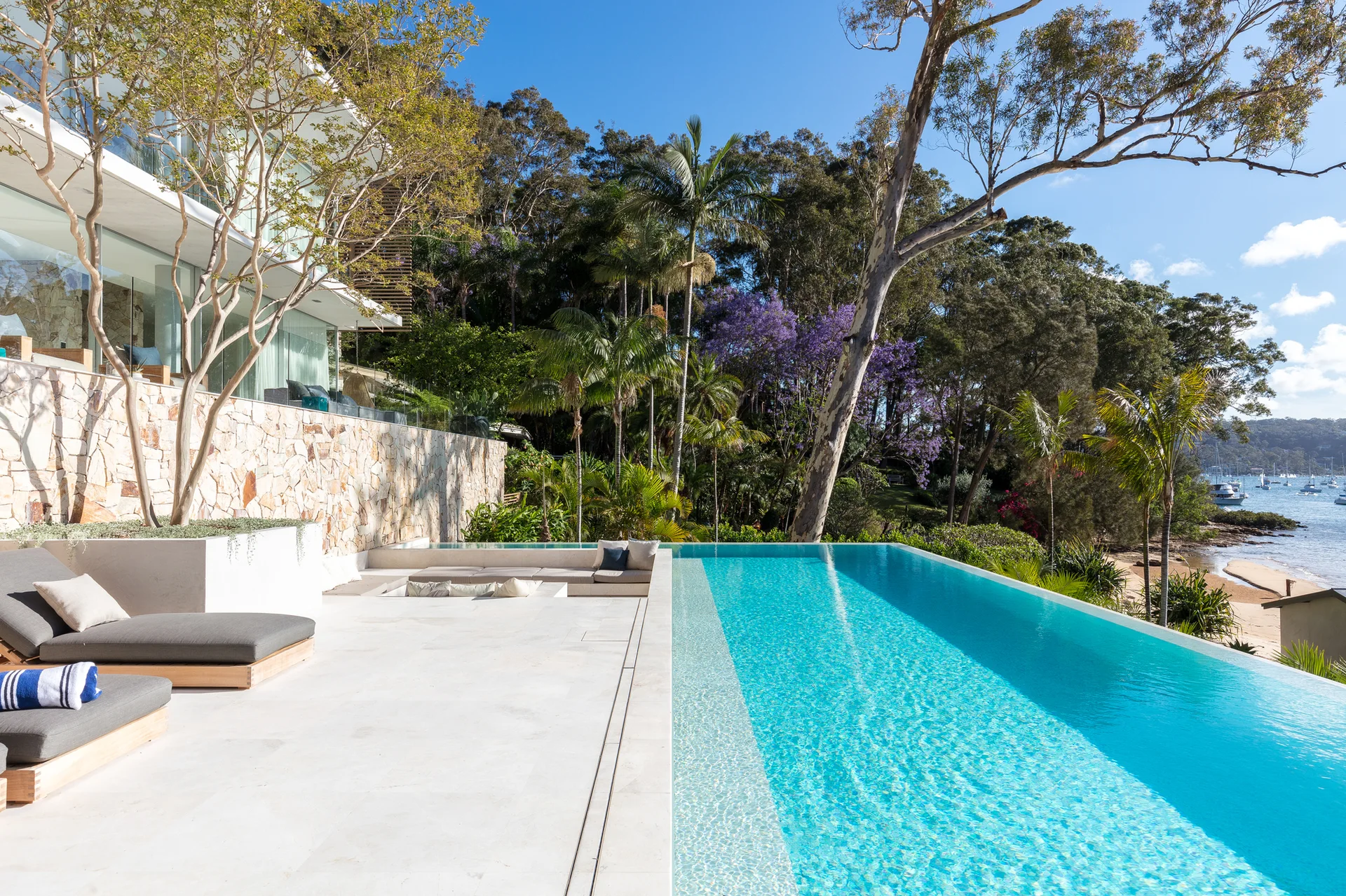 An outdoor terrace features a turquoise infinity pool, grey lounge chairs on off-white pavers, and a stacked stone retaining wall.