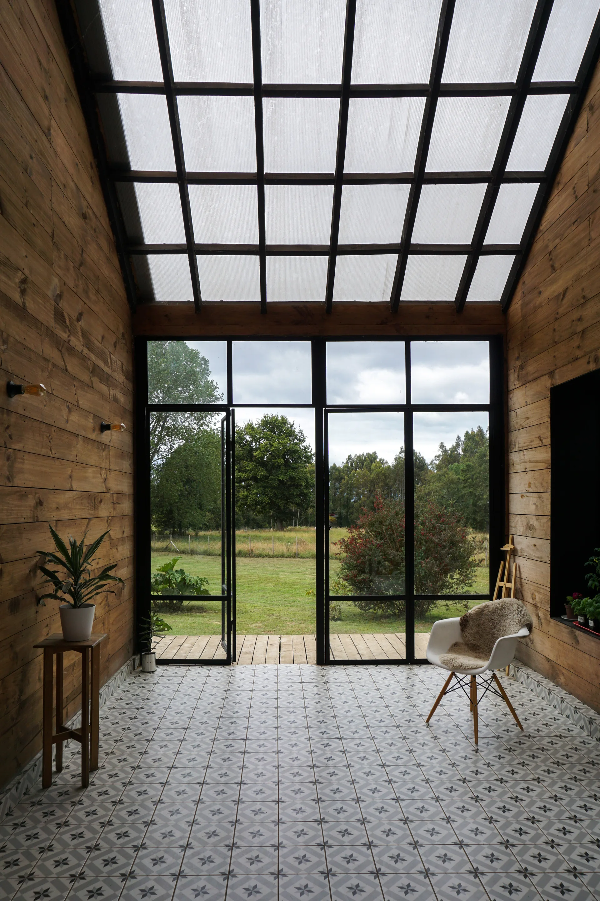 A rustic sunroom featuring horizontal wood plank walls, geometric star-patterned floor tiles, and large black metal glass doors leading to a garden.