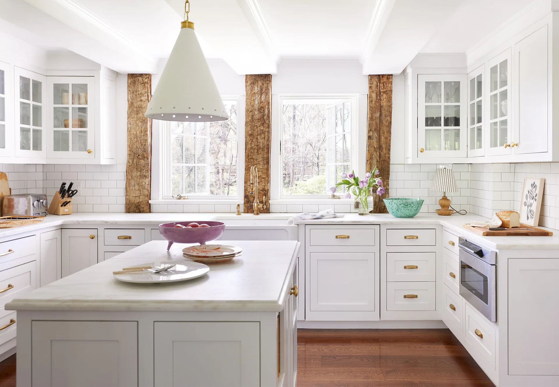 A bright white kitchen featuring shaker cabinets, a marble-topped island, an apron sink, and rough-hewn wooden posts flanking two windows.