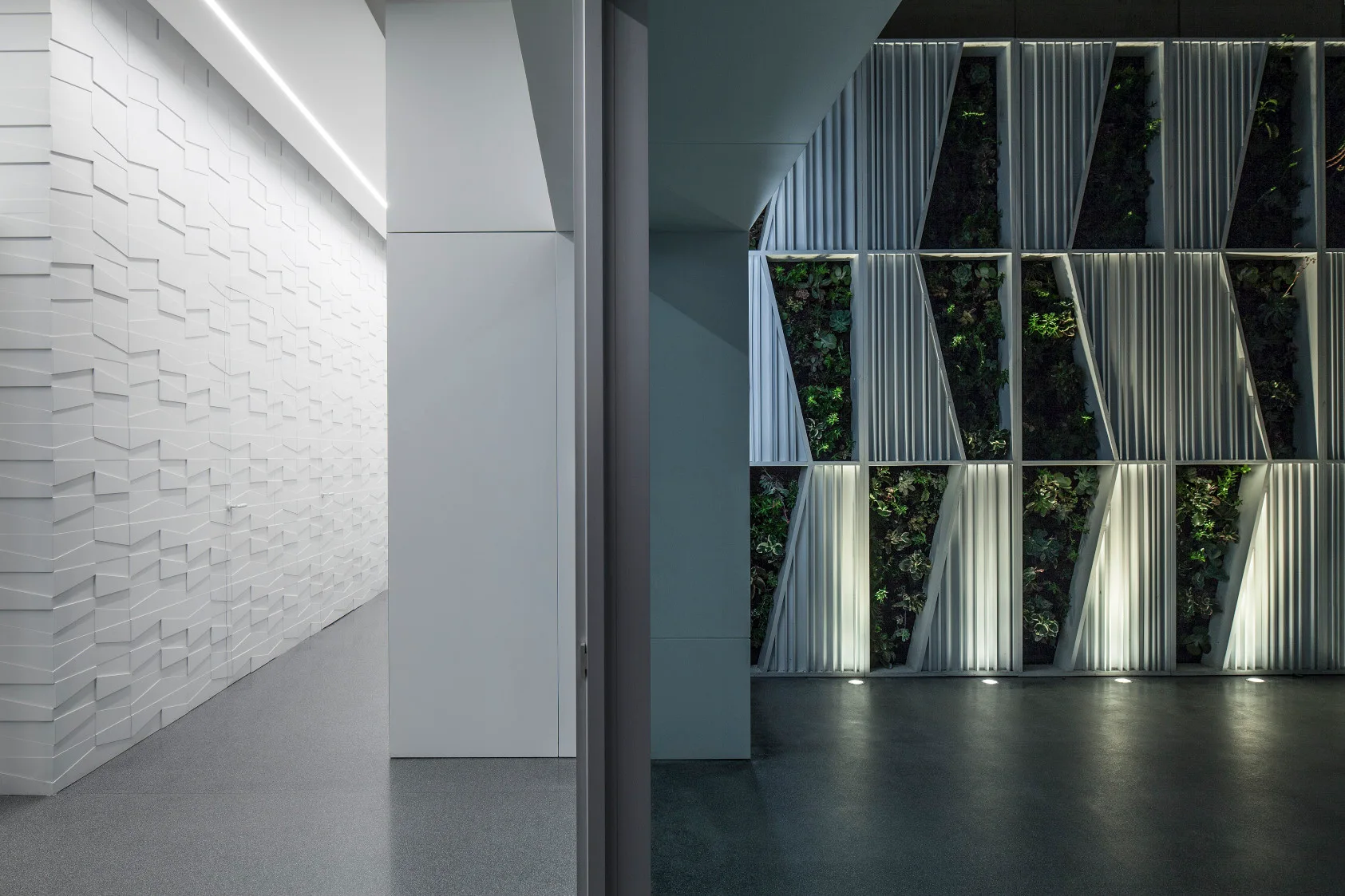 A minimalist corridor features a white geometric 3D textured wall opposite a structured vertical garden panel illuminated by floor uplights.