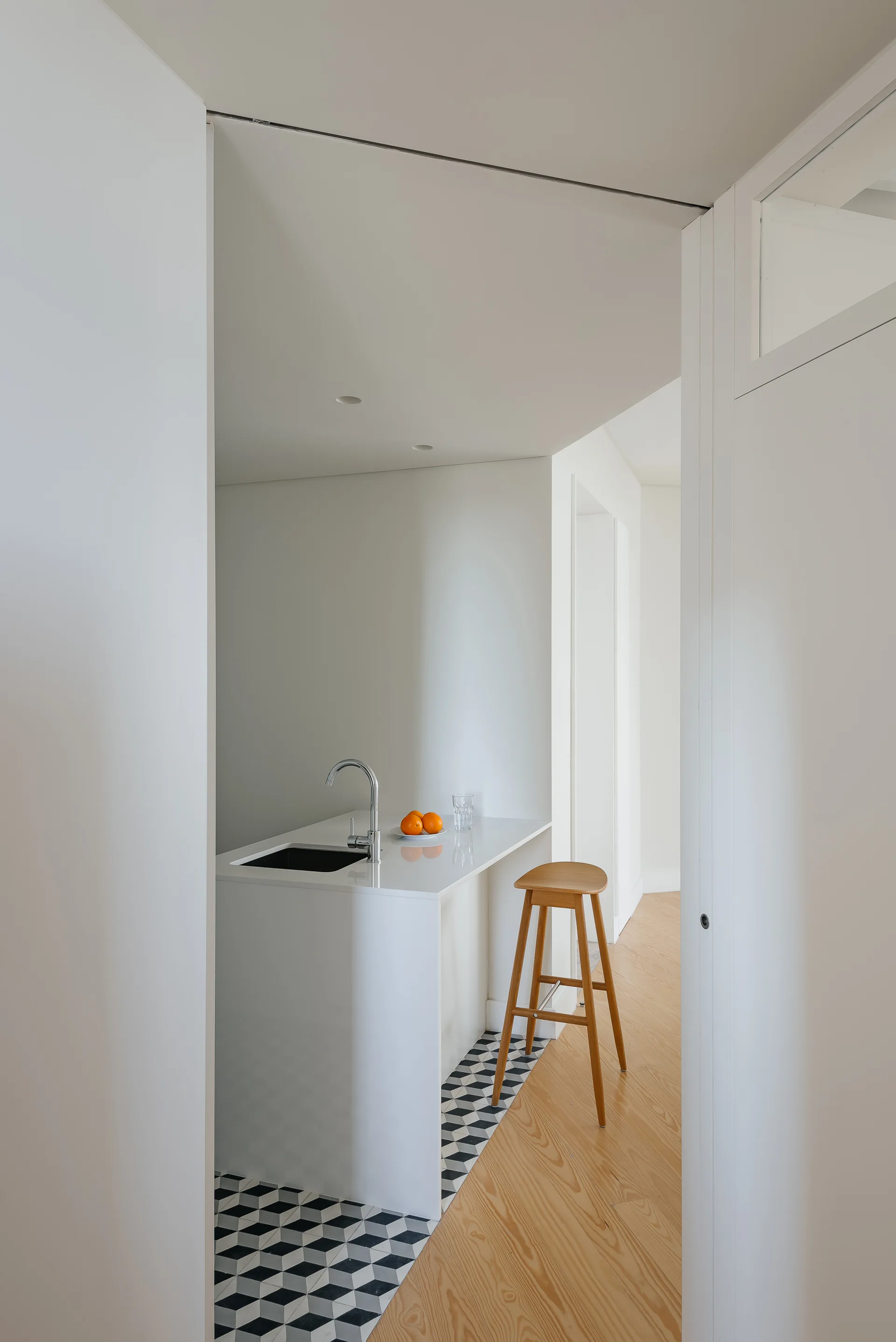 A minimalist white kitchen island with a chrome faucet stands on geometric tiles next to a wooden stool on hardwood flooring.
