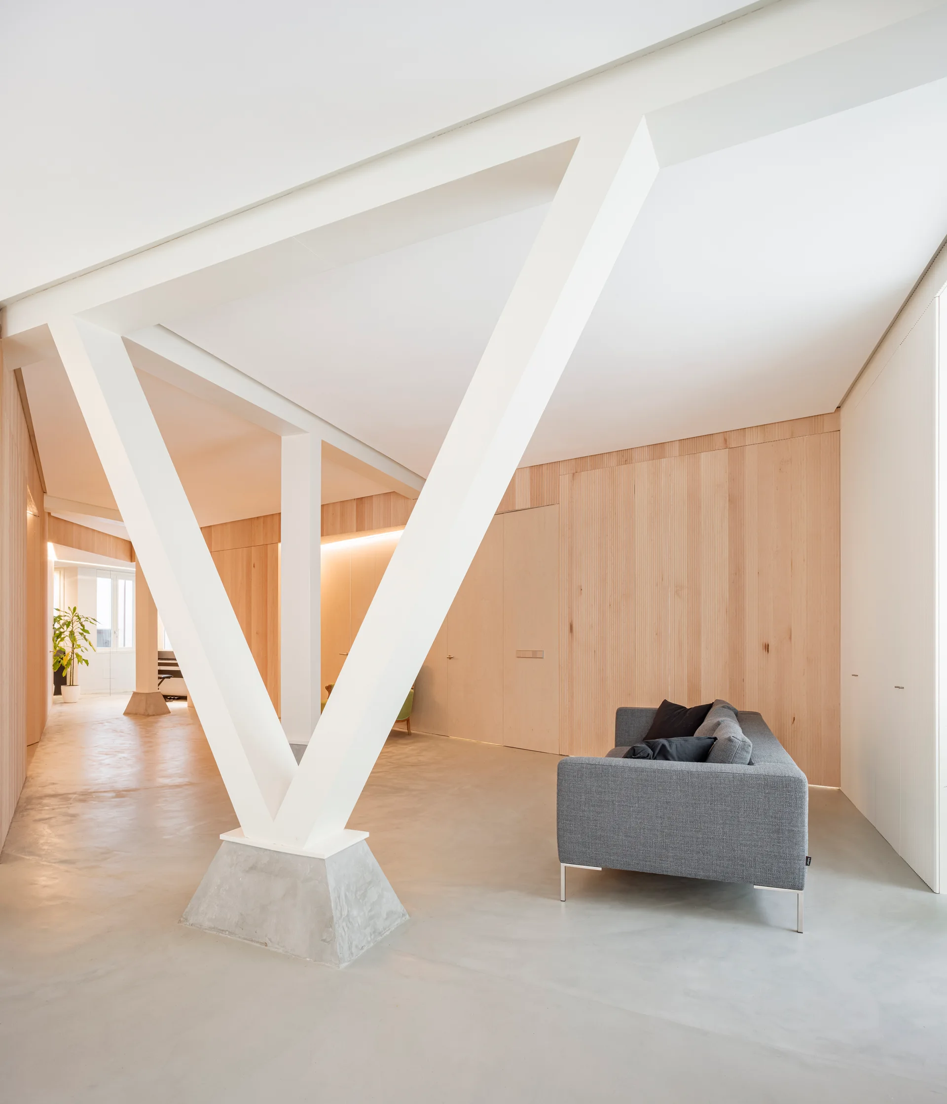 A minimalist living room featuring a grey sofa, light wood paneled walls, and a distinctive white V-shaped structural column.