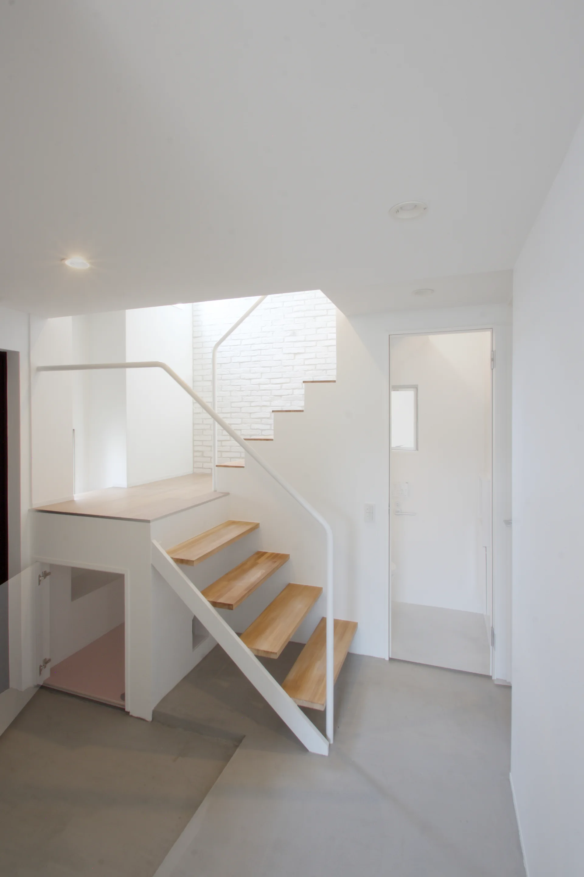 A modern open-tread staircase with light wood steps and a white metal handrail stands in a minimalist hallway with white walls and a grey floor.