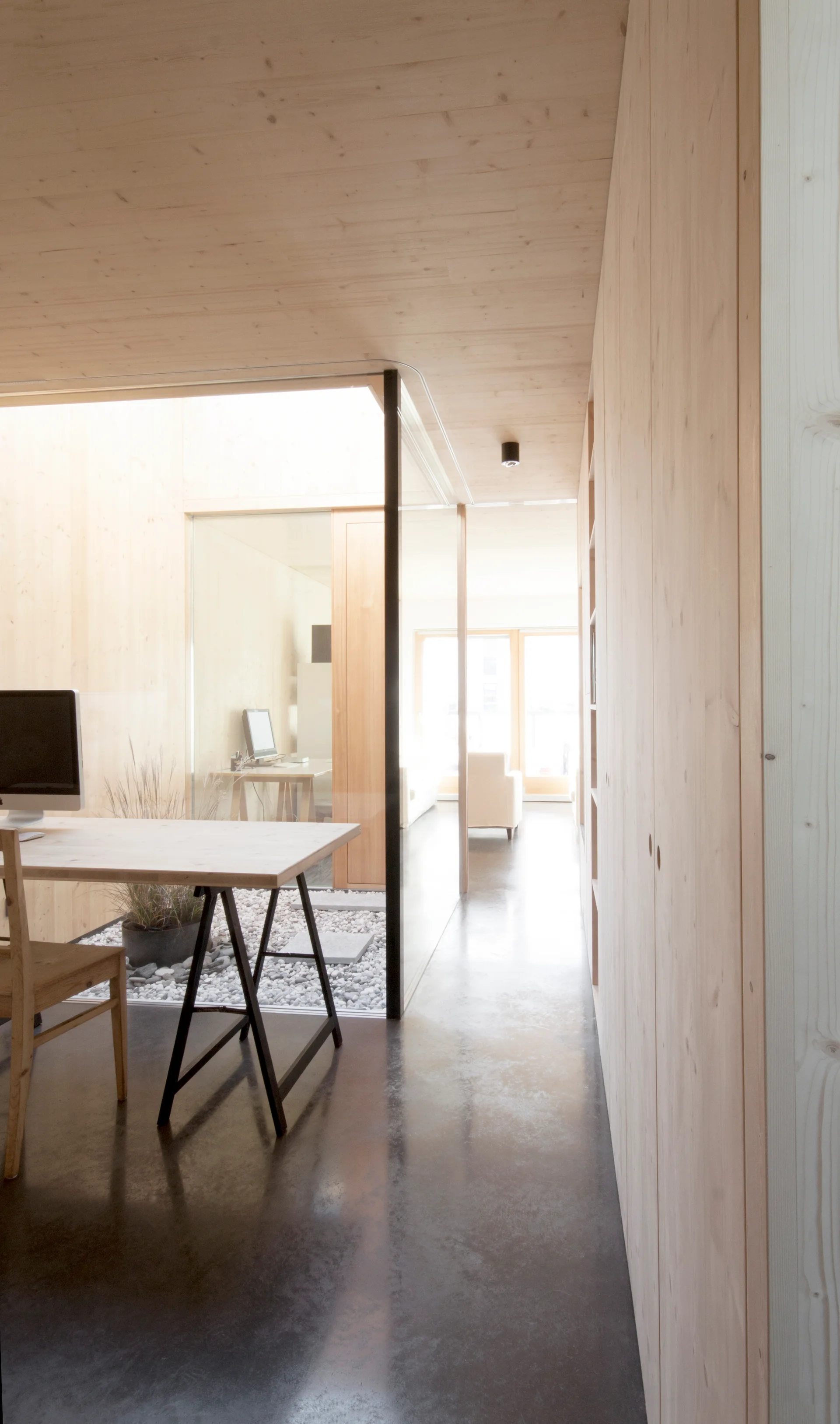 A minimalist home office features light wood walls and ceiling, a dark concrete floor, and a glass-walled indoor courtyard with gravel.