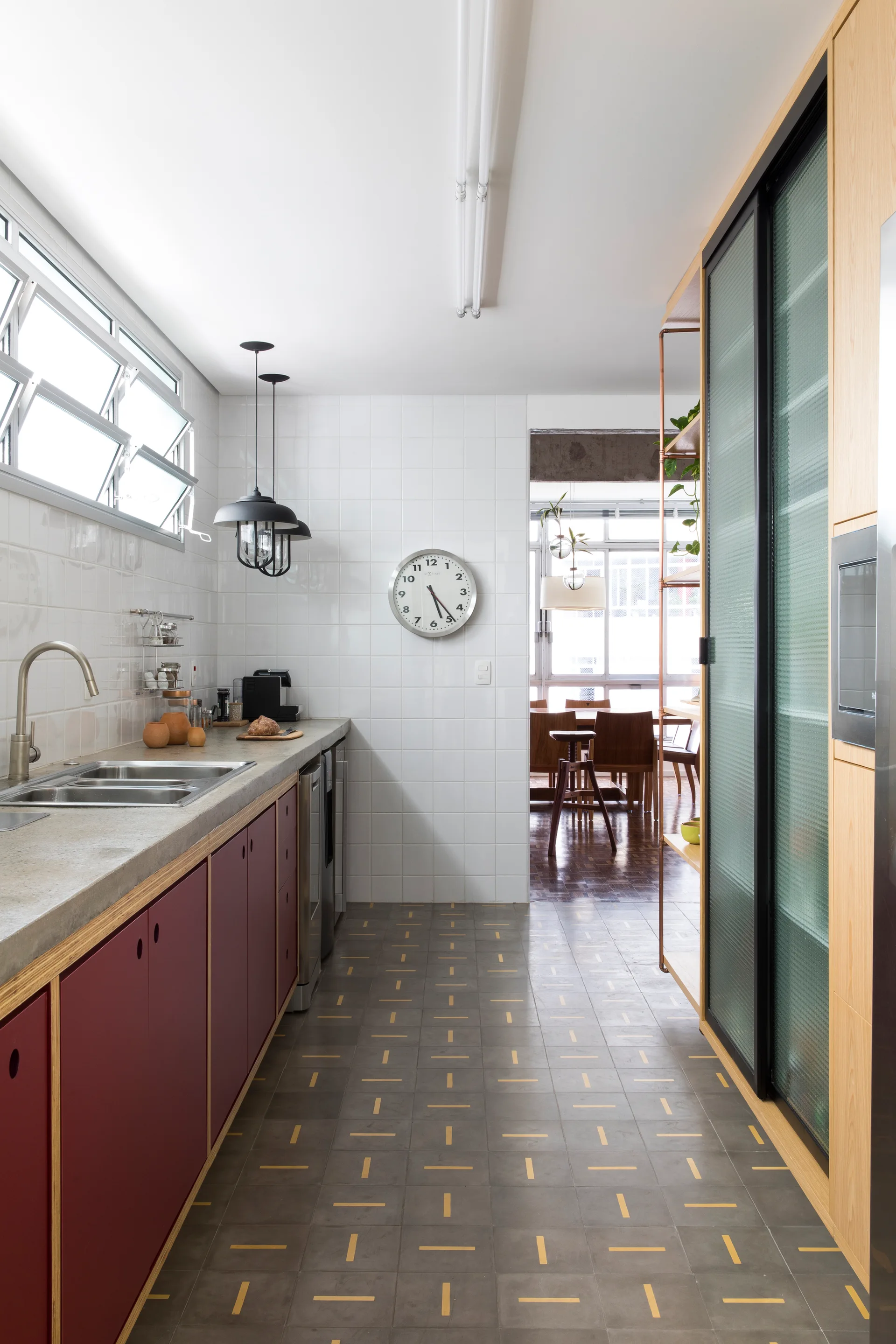 A galley kitchen features maroon base cabinets with exposed plywood edges, grey concrete countertops, patterned grey floor tiles, and white wall tiles.
