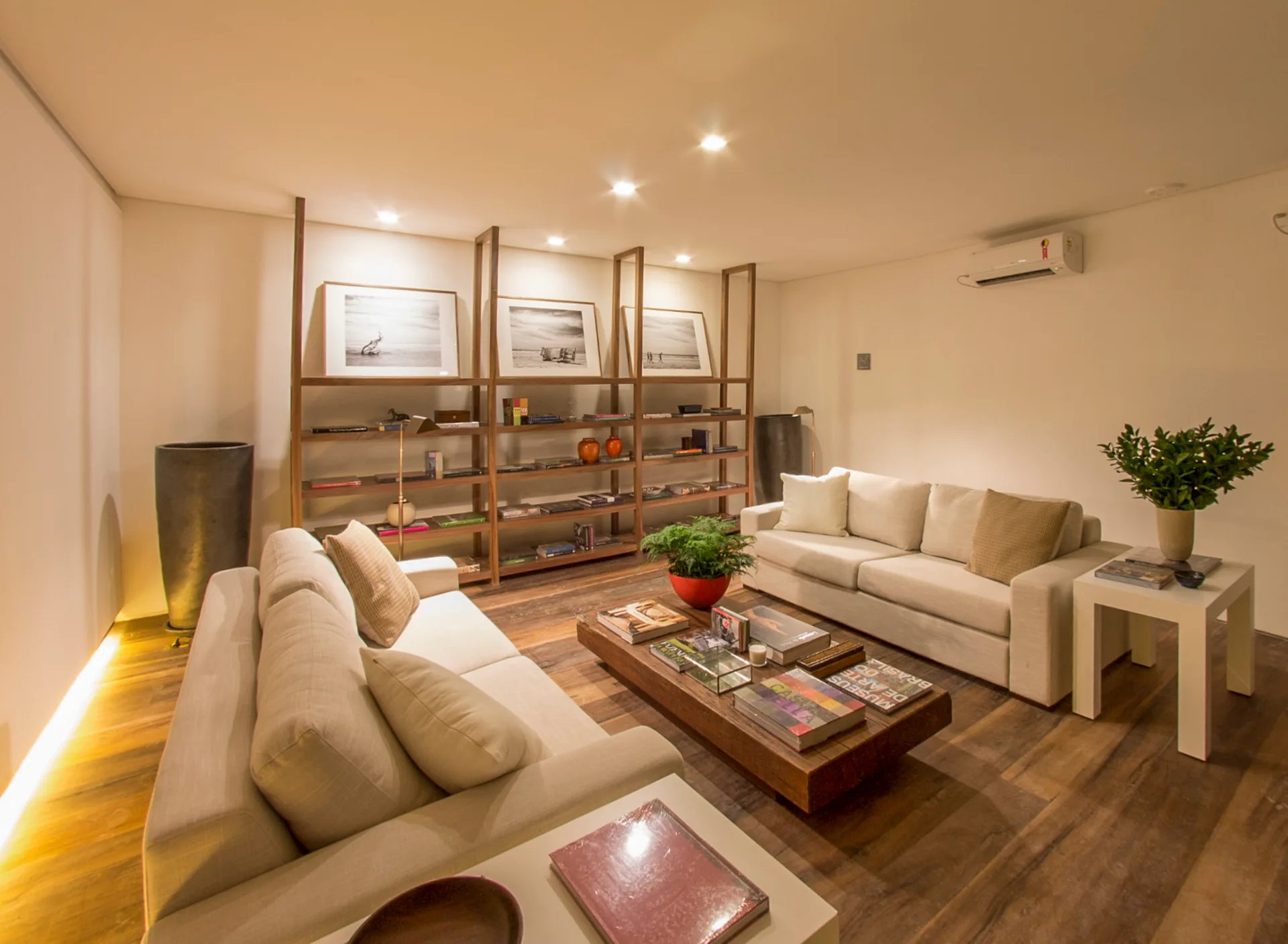 Two beige sofas face each other over a low wood coffee table in a room featuring a large wooden open shelving unit and warm recessed lighting.