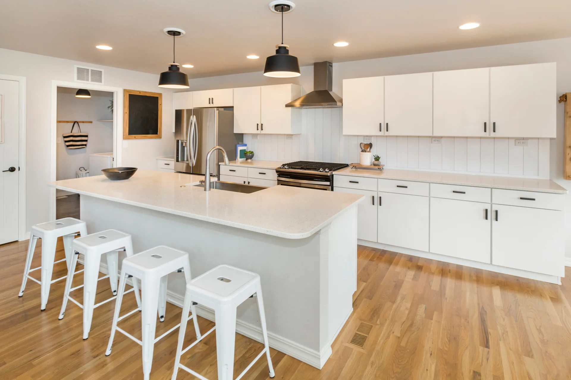 A bright kitchen features white cabinetry, a central island with white metal stools, a vertical plank backsplash, and black dome pendant lights.