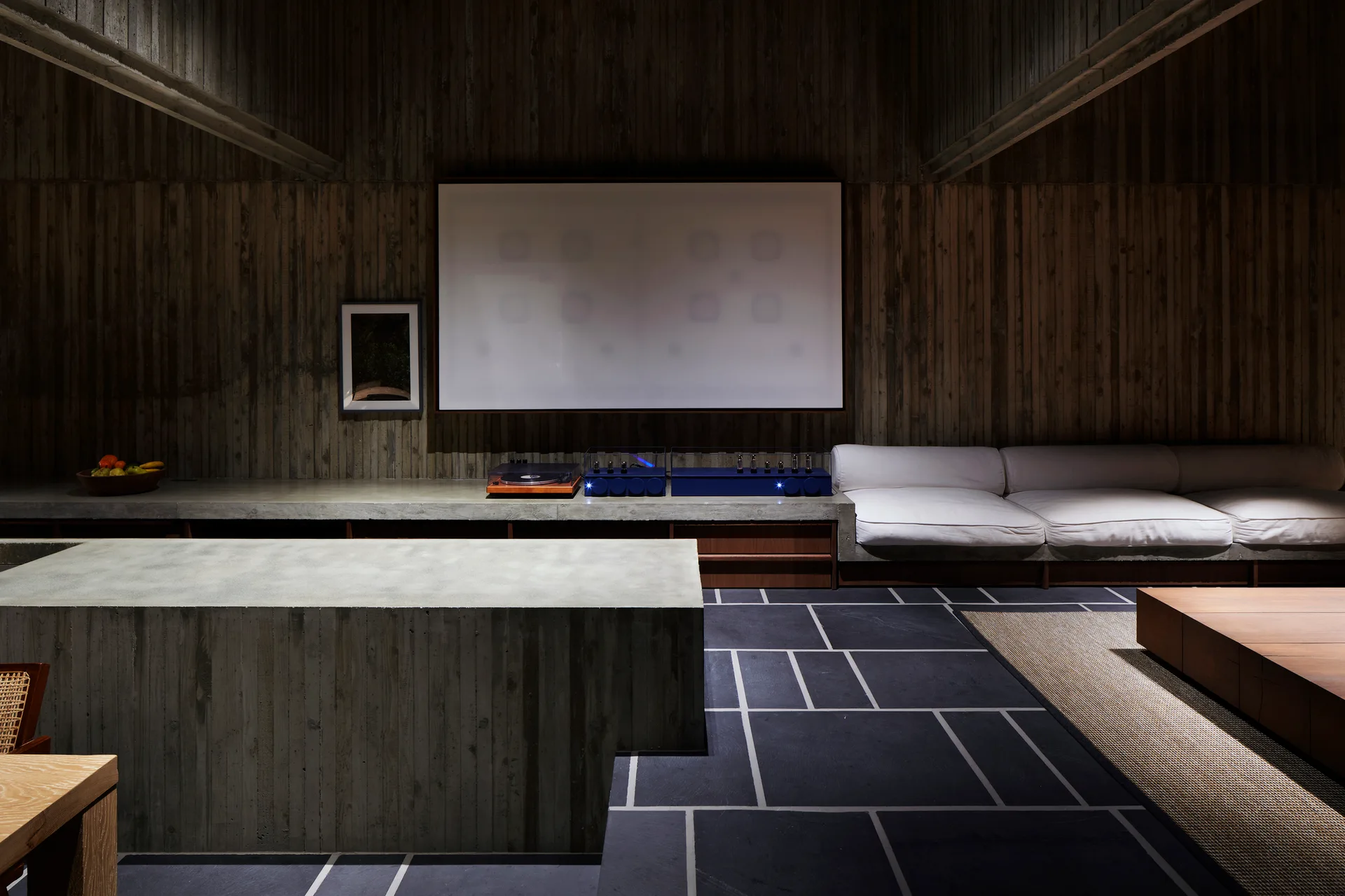 A minimalist room with textured, board-formed concrete walls, a low white modular sofa, a concrete block island, and dark stone tile flooring.