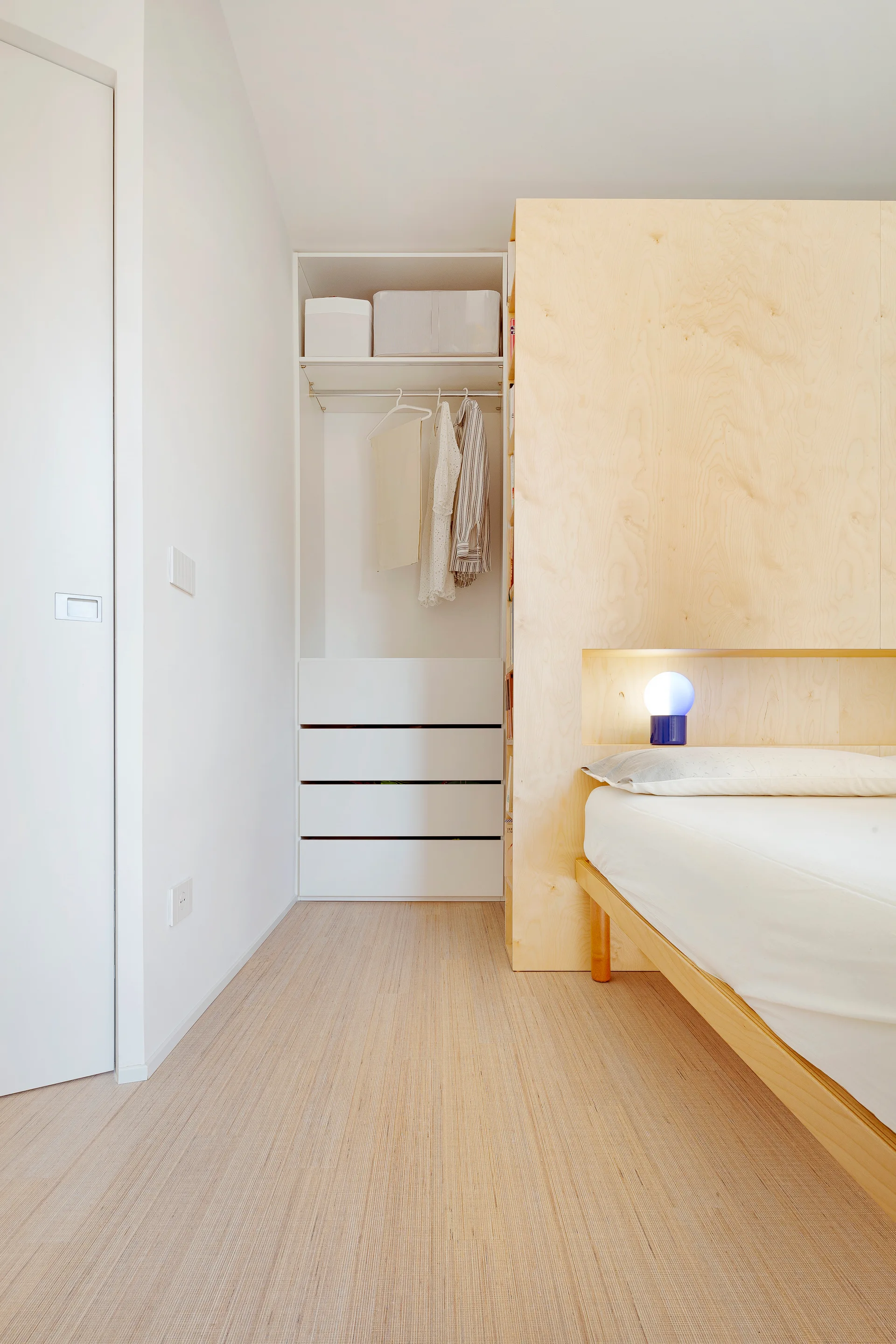 A minimalist bedroom features a light wood headboard unit that acts as a room divider next to an open closet with white drawers.
