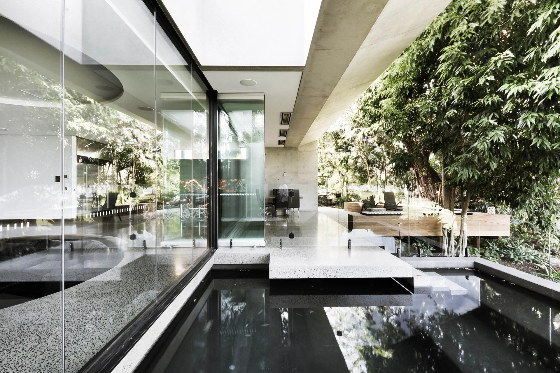 A modern architectural patio featuring floor-to-ceiling glass walls next to a dark reflection pool with a floating concrete walkway.