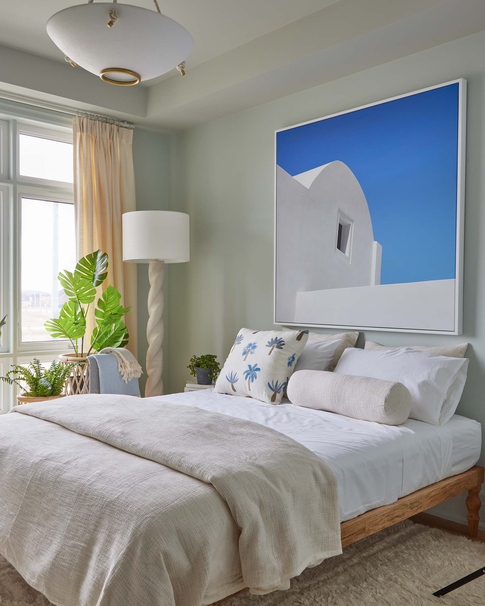 A bedroom with sage green walls, a wooden bed with white linens, a large blue and white framed photo, and a twisted floor lamp.