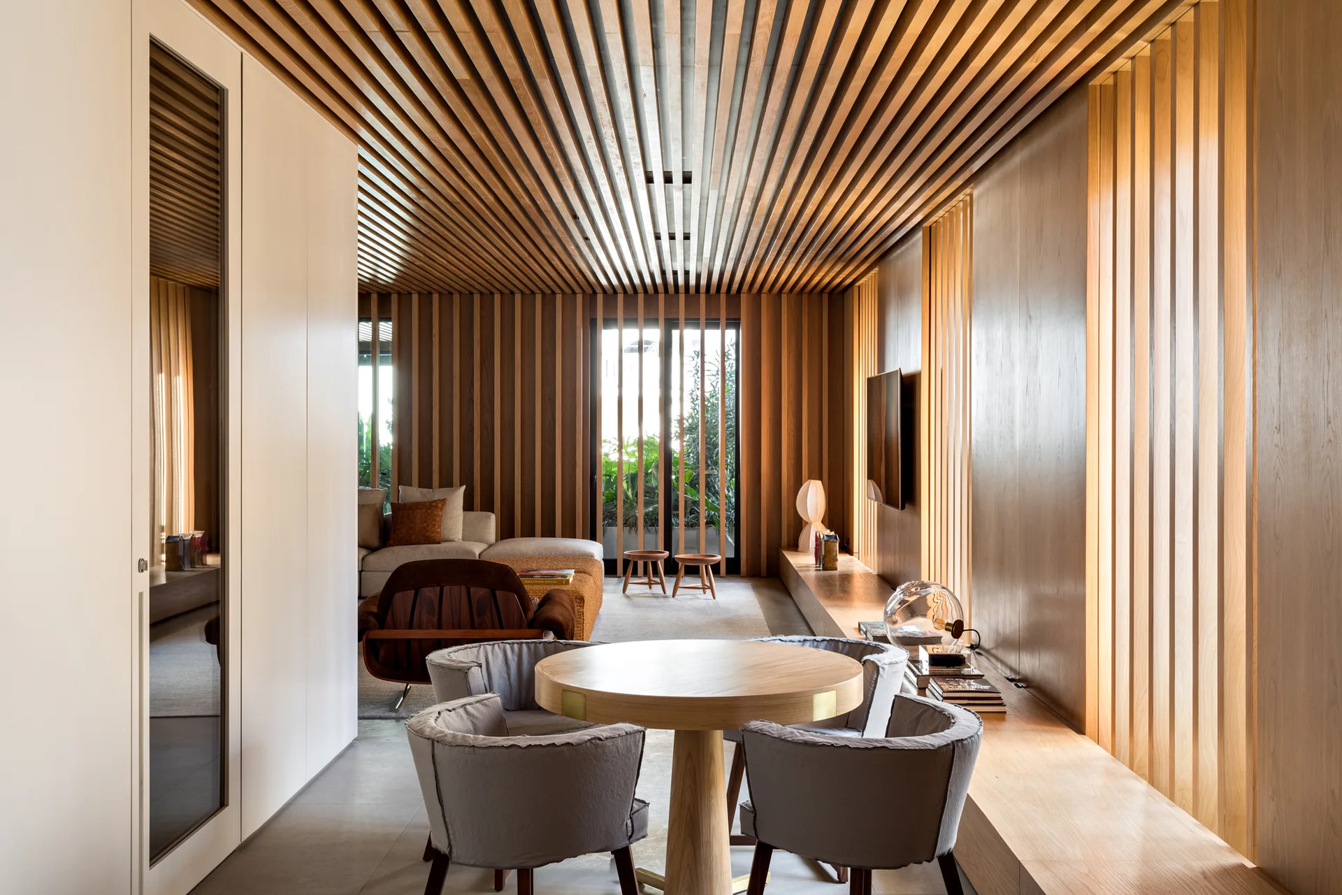 A modern open-plan dining space with a round light wood table and grey chairs set against vertical wood slat cladding on the wall and ceiling.