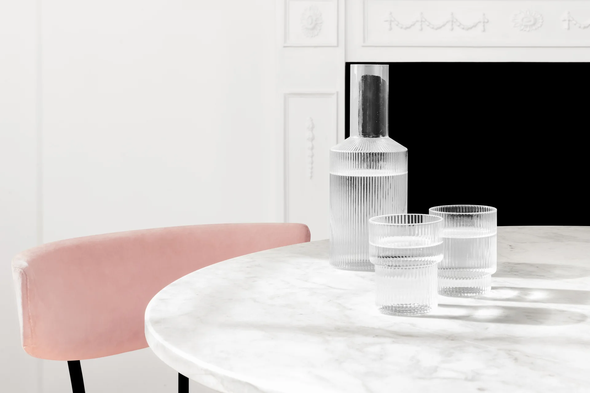 A ribbed glass carafe and tumblers rest on a round marble dining table next to a pale pink upholstered chair in front of a white fireplace.