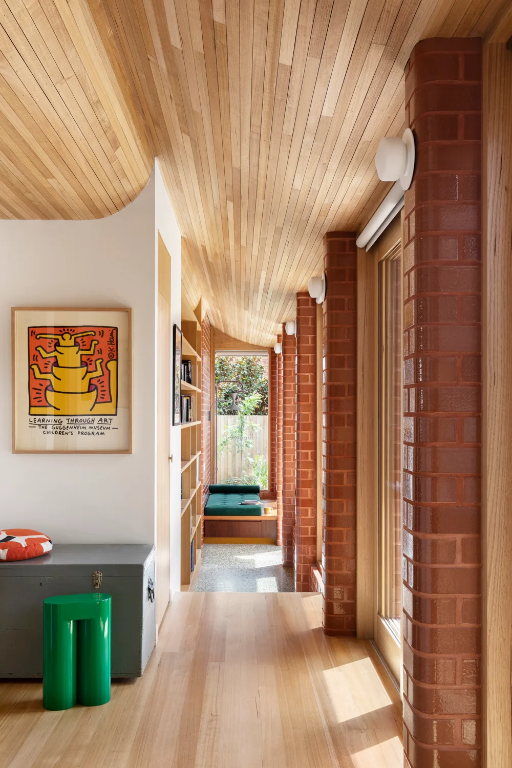 A sunlit hallway featuring a curved wood-planked ceiling, glazed brick columns, built-in wooden bookshelves, and a far window seat.