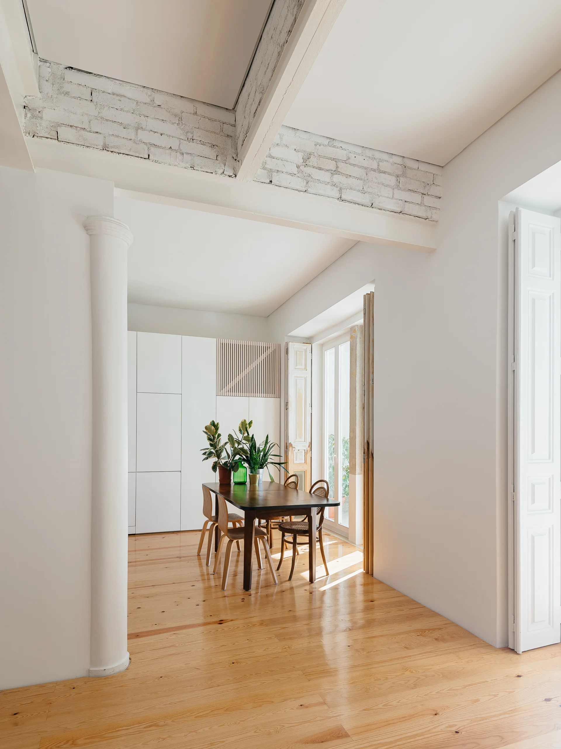 A minimalist dining room features a dark wood table and chairs on glossy wood floors, with white built-in cabinets and an exposed brick ceiling beam.