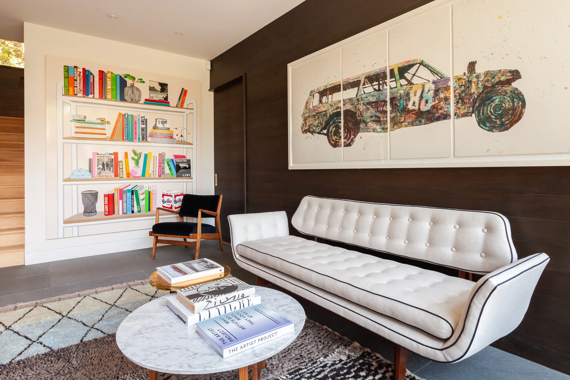 A mid-century modern white tufted sofa sits against dark horizontal wood paneling in a living room with a round marble coffee table and bookshelf mural.