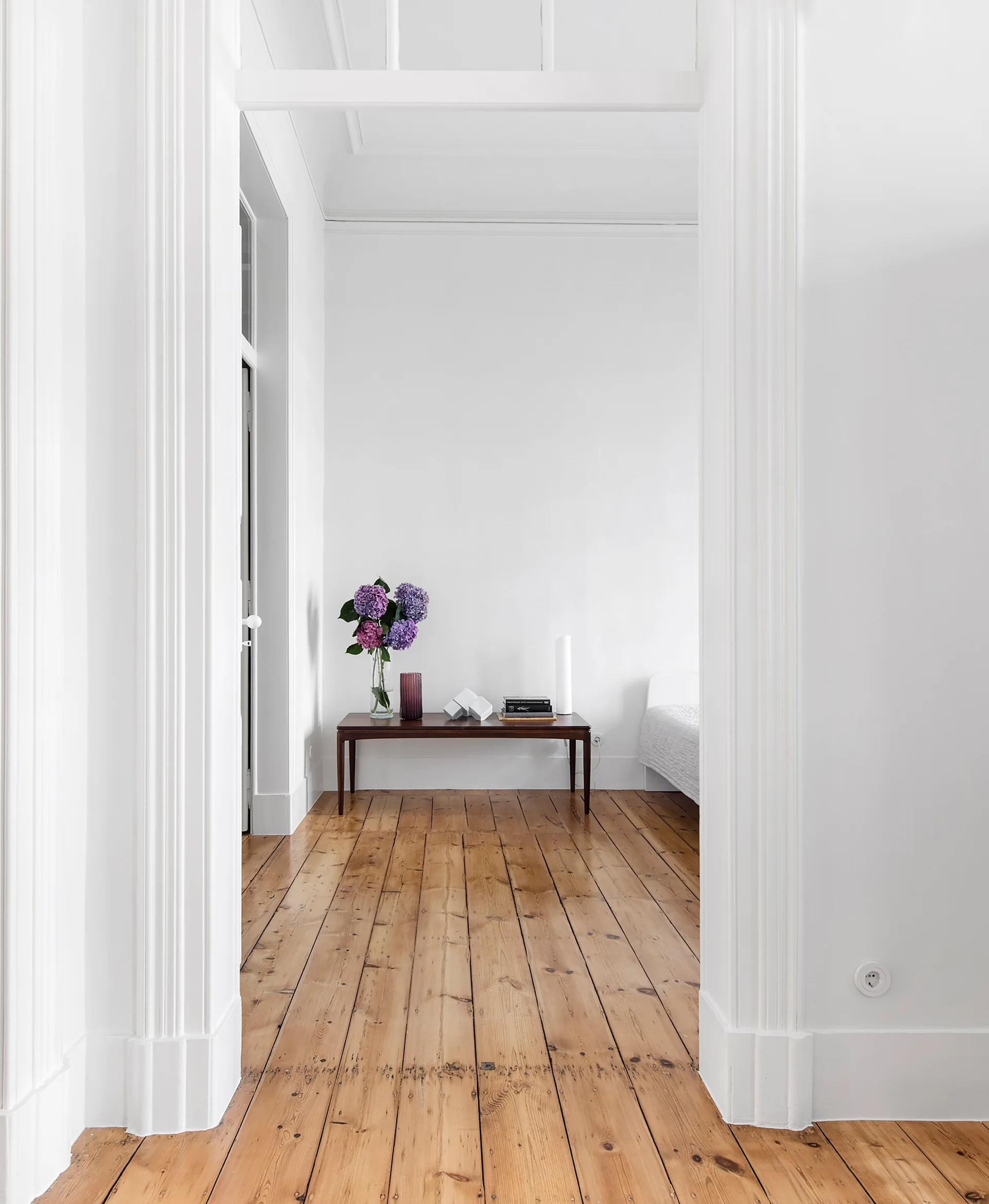 A view through a wide white door frame into a bright room featuring natural wood plank floors and a low dark wood console table decorated with flowers.