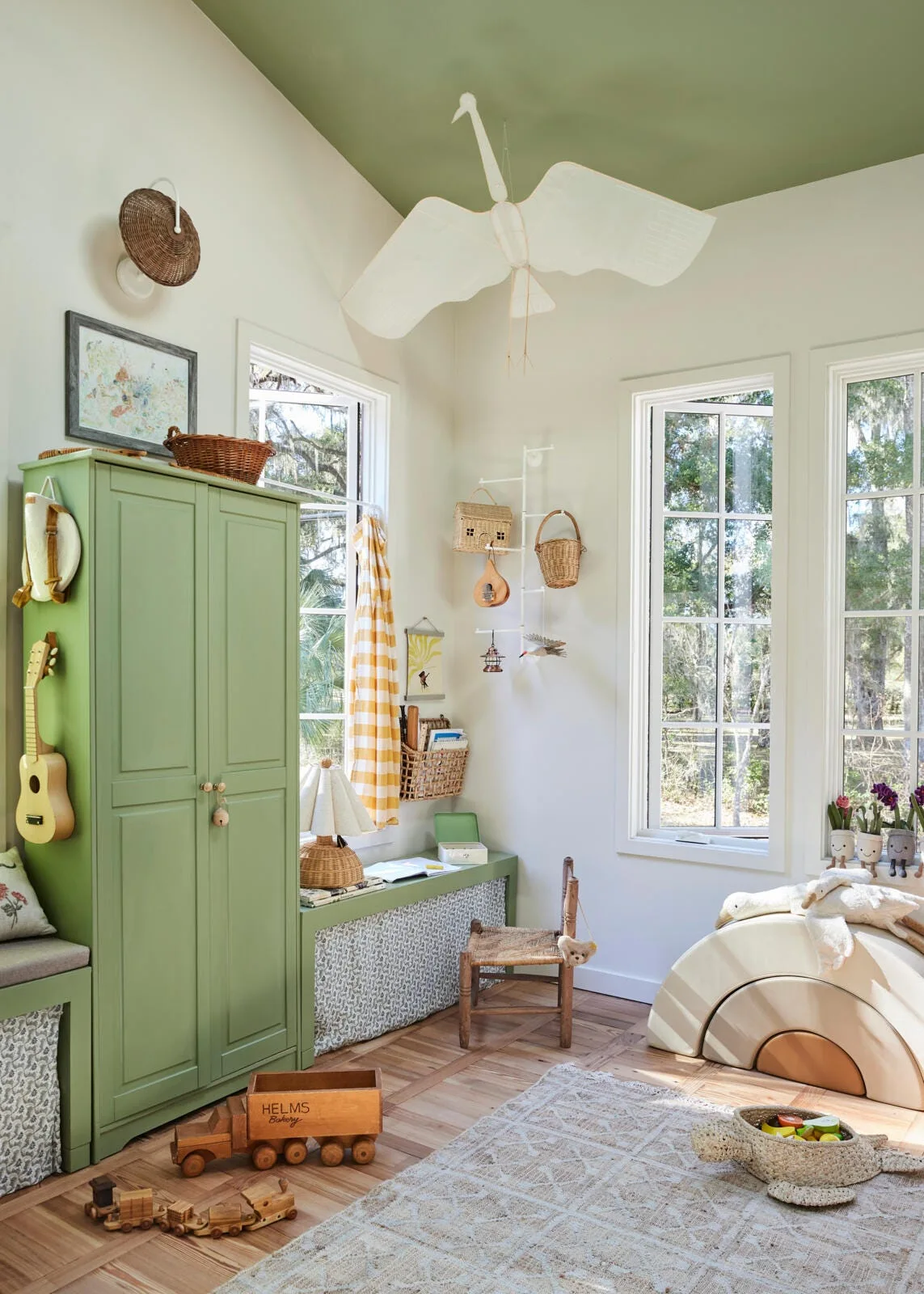 A bright playroom features a sage green wardrobe, a matching painted ceiling, and a large white fabric bird mobile hanging over wooden toys.