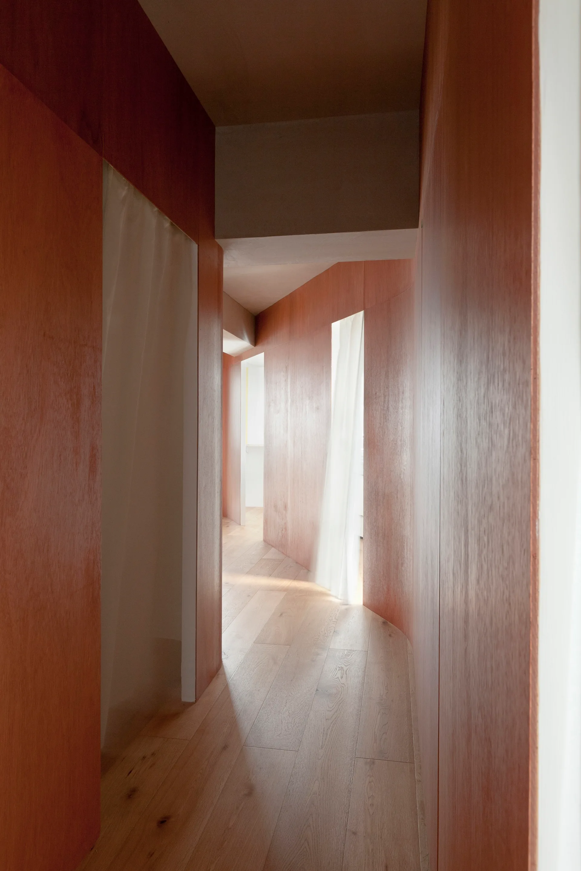 A narrow hallway with warm red-toned wood-paneled walls, light wood plank flooring, and soft white curtains hanging in the doorways.