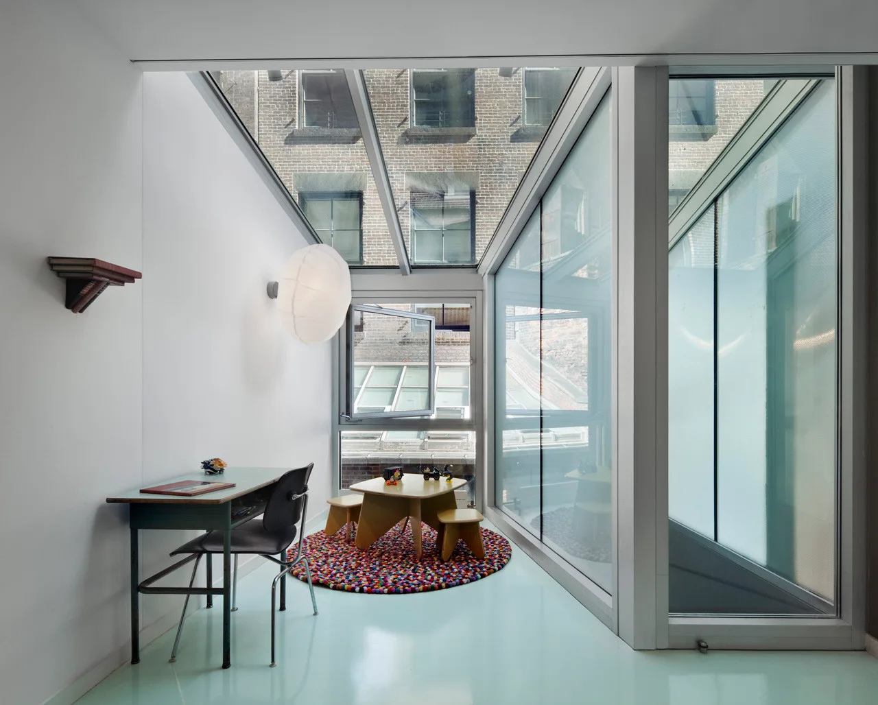 A modern playroom in a glass sunroom featuring mint green floors, child-scale furniture, and a colorful felt-ball rug.
