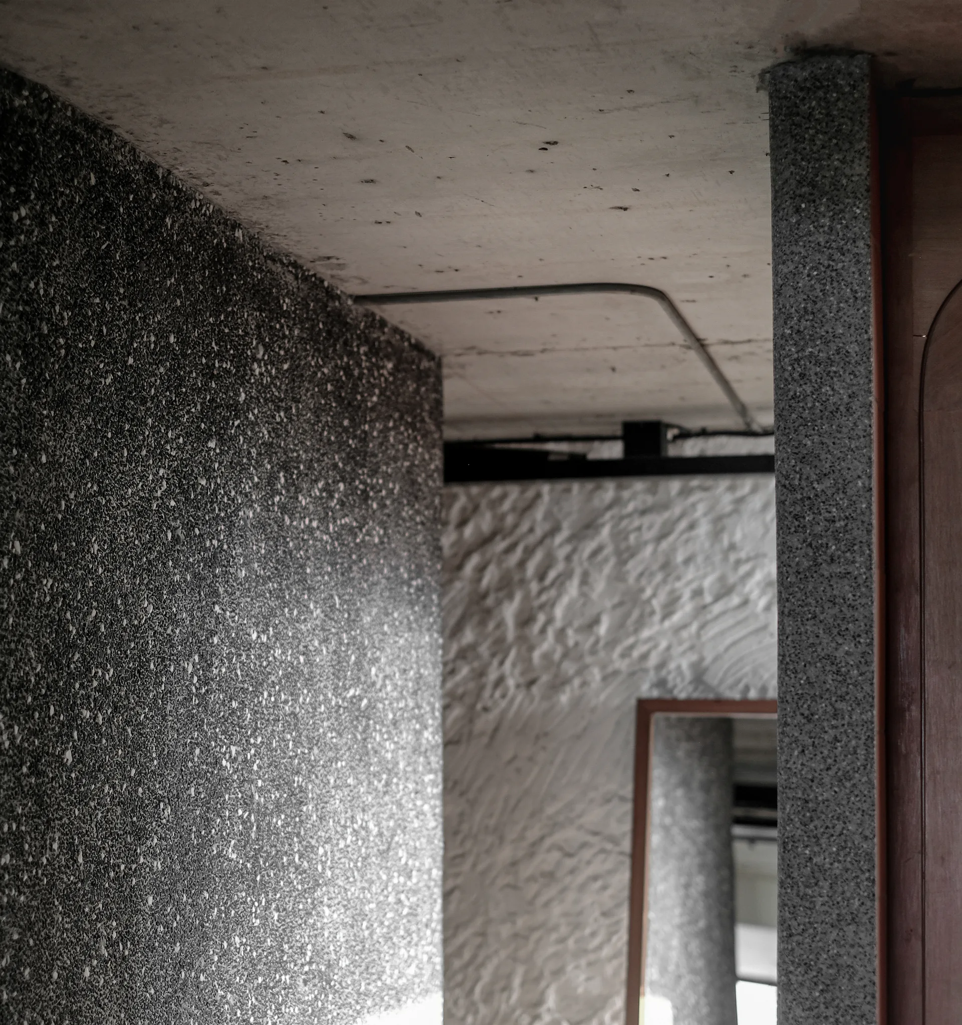 A close-up view of an interior corridor with a raw concrete ceiling, a dark speckled wall, and a textured white plaster wall in the background.