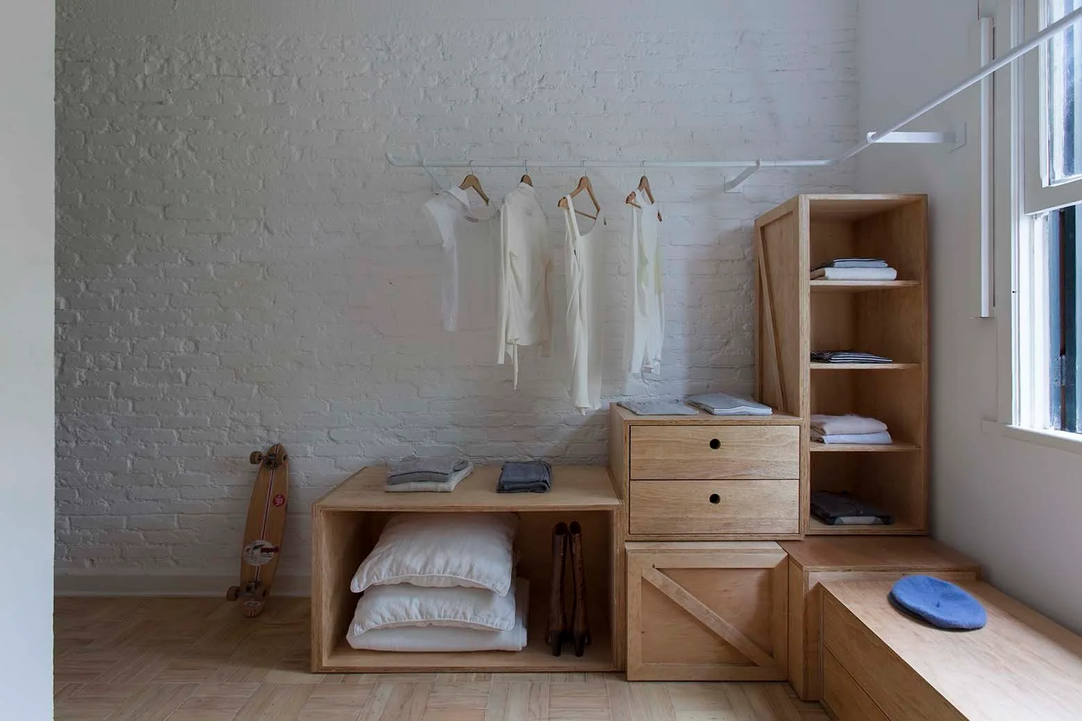 A modular light wood closet system with drawers and open shelves is arranged against a painted white brick wall next to a clothing rack.