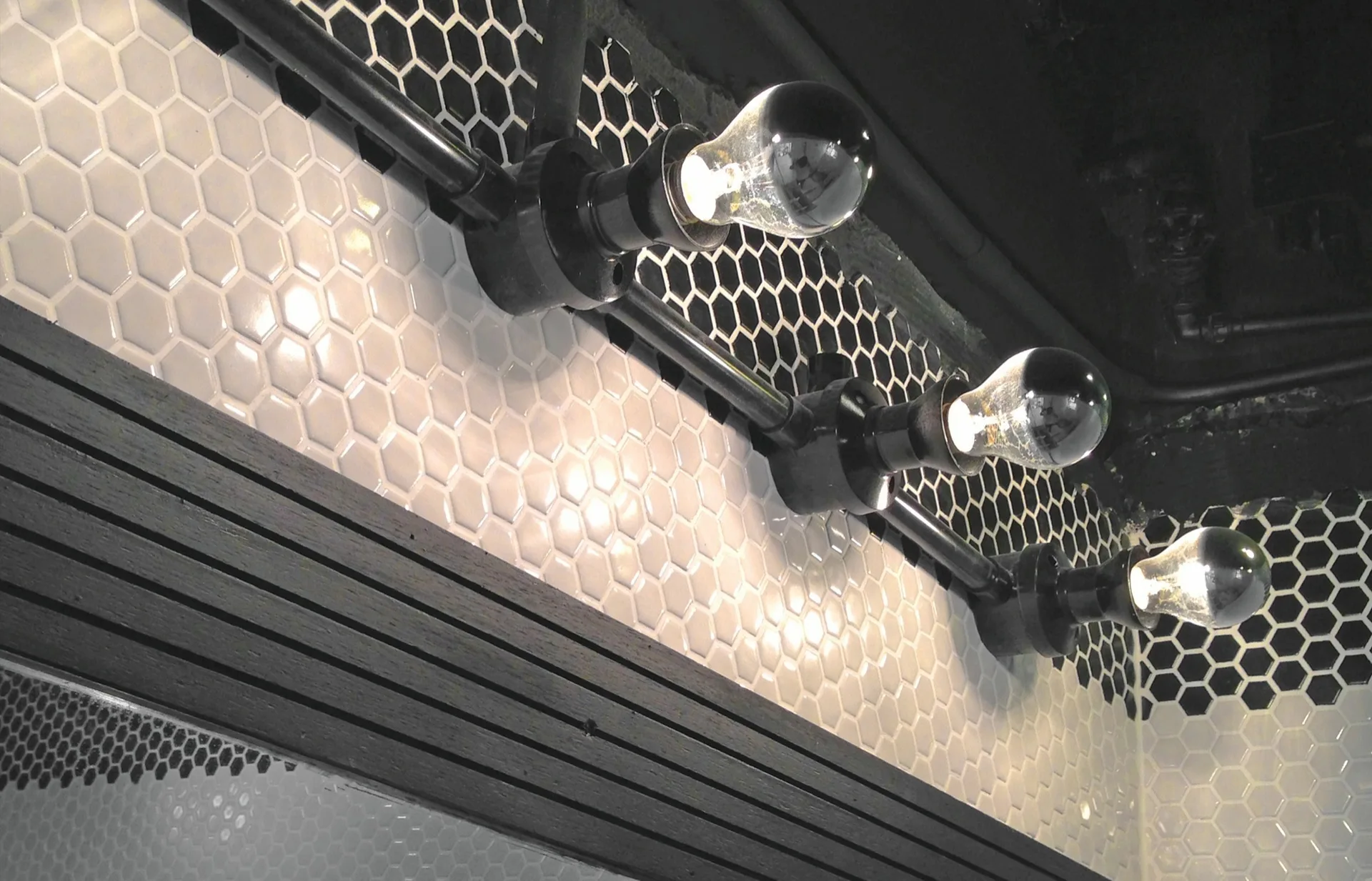 An industrial black vanity light fixture with three exposed bulbs mounted on a white and black hexagon tile wall.