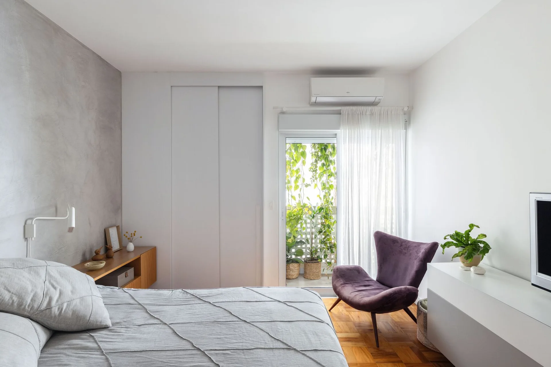 A minimalist bedroom featuring a grey bed against a concrete accent wall, a purple armchair on parquet flooring, and a plant-filled balcony.