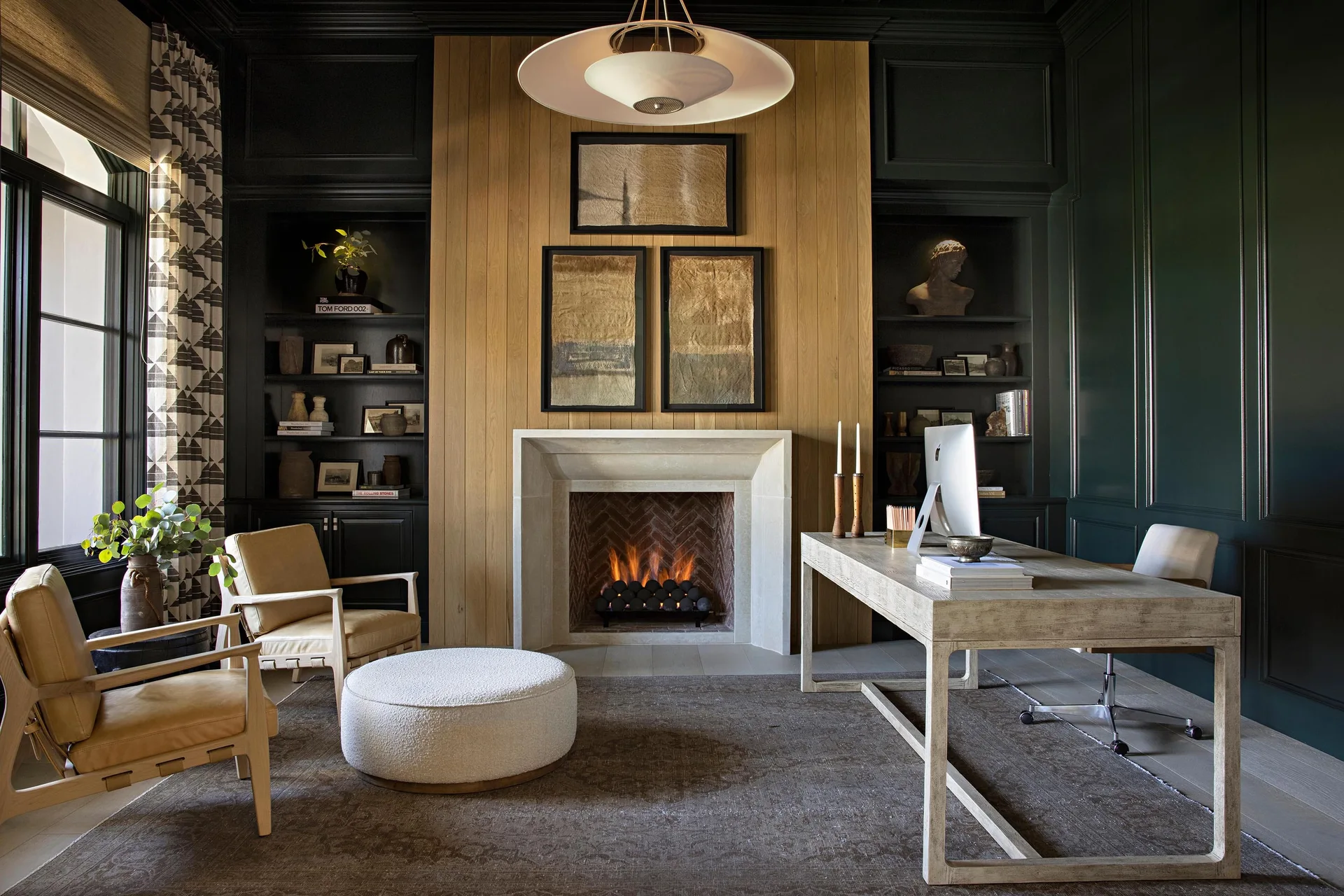 A dark green home office features a central stone fireplace with vertical wood slat paneling, flanked by dark built-in bookshelves and a wooden desk.