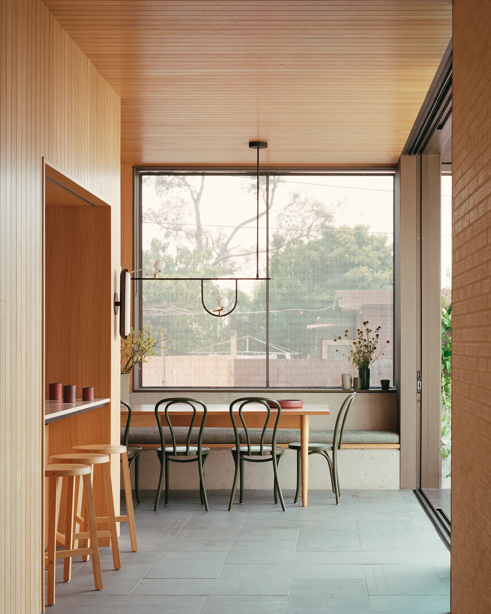 A modern dining area features light wood wall paneling, a matching dining table with dark green bentwood chairs, and a large window.