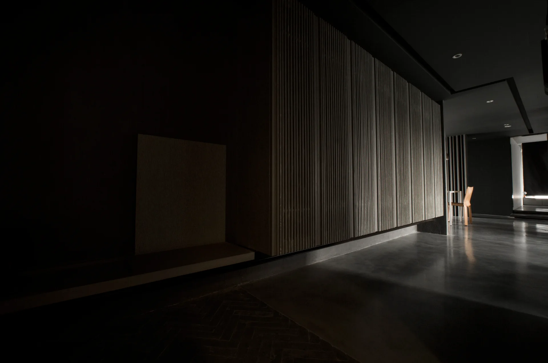 A dark modern interior hallway featuring a large light wood slatted wall panel, a polished concrete floor, and a wooden chair in the distant background.