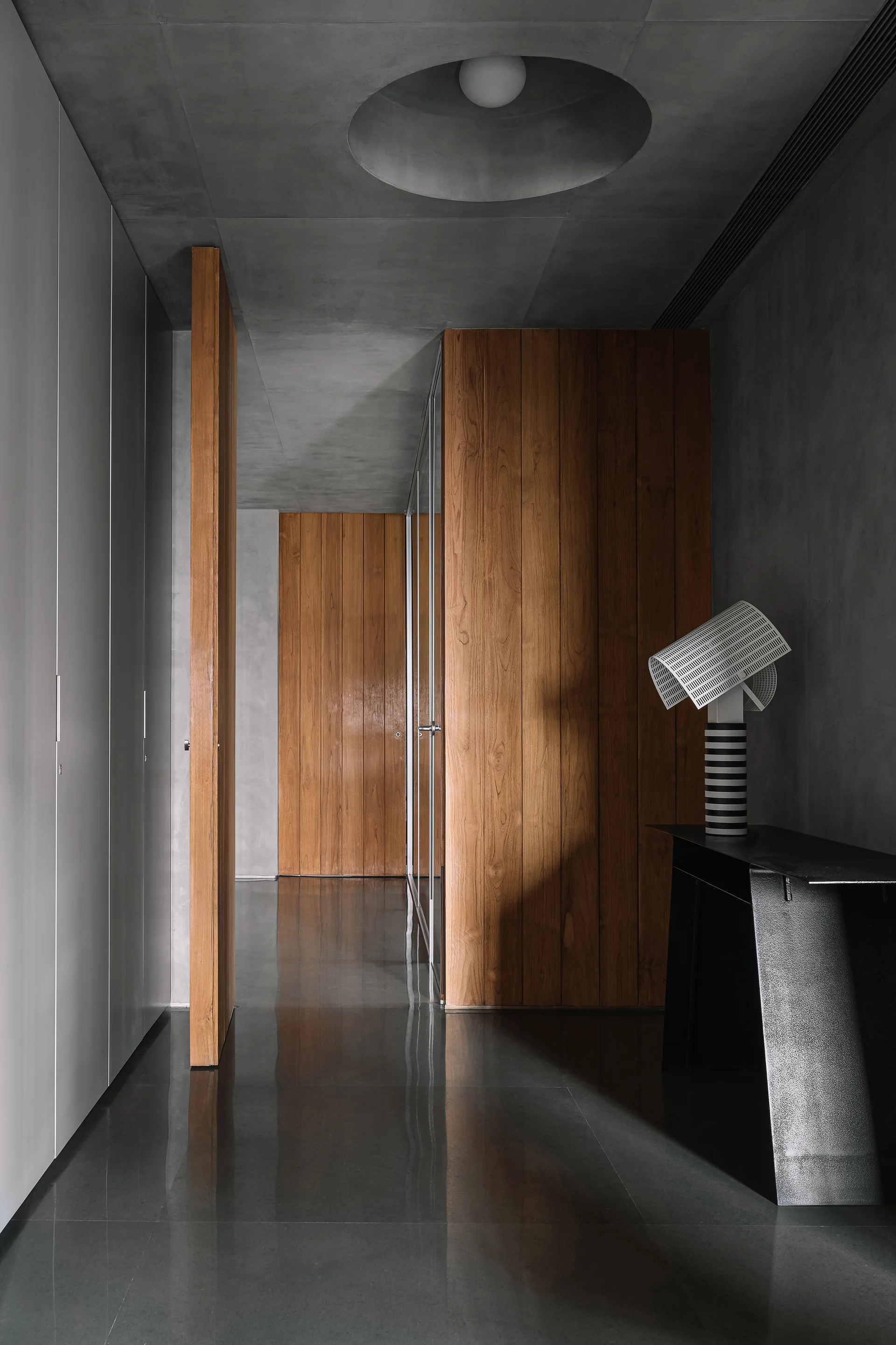 A contemporary hallway featuring vertical wood panels, a smooth concrete ceiling with a circular recessed light, a black console table, and a reflective floor.