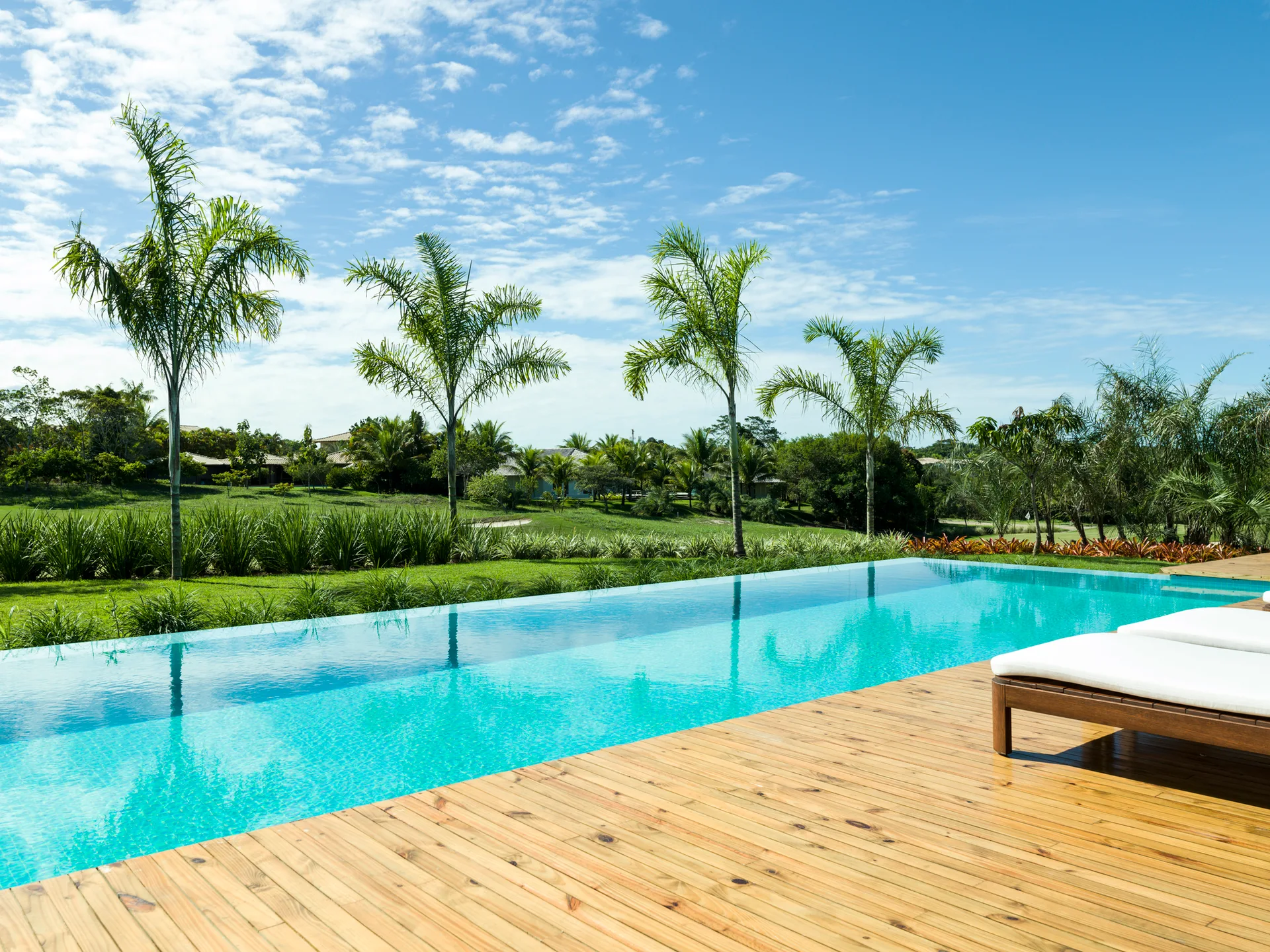 An infinity edge swimming pool with turquoise water sits next to a light wood deck with white lounge chairs overlooking a garden with palm trees.