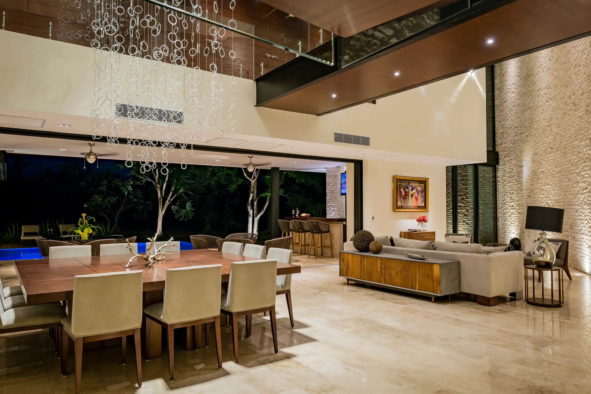 A spacious open-concept dining and living area features a large wooden dining table, polished stone flooring, and wide glass doors opening to a pool.