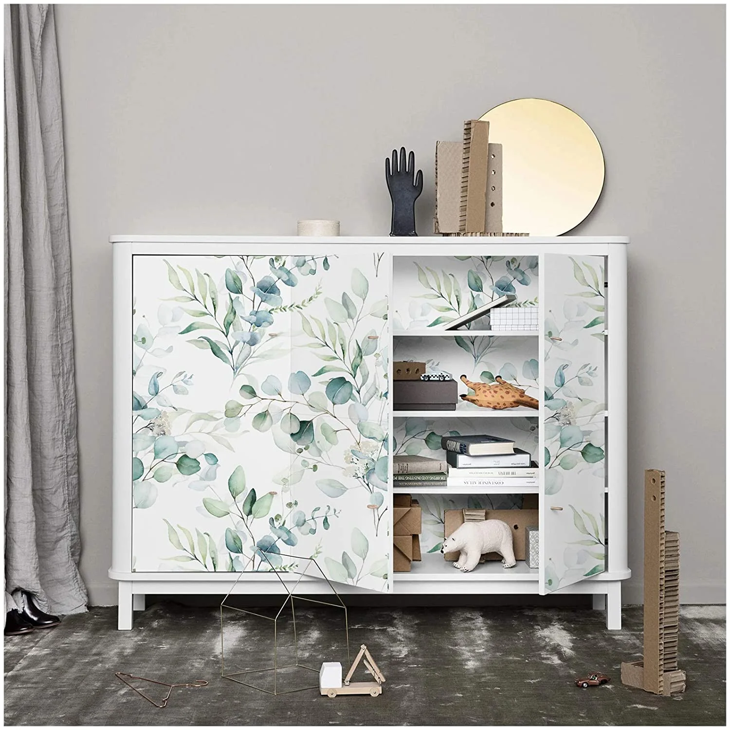 A white wooden cabinet featuring green botanical-patterned doors stands open against a gray wall, showing toys and books on the inner shelves.