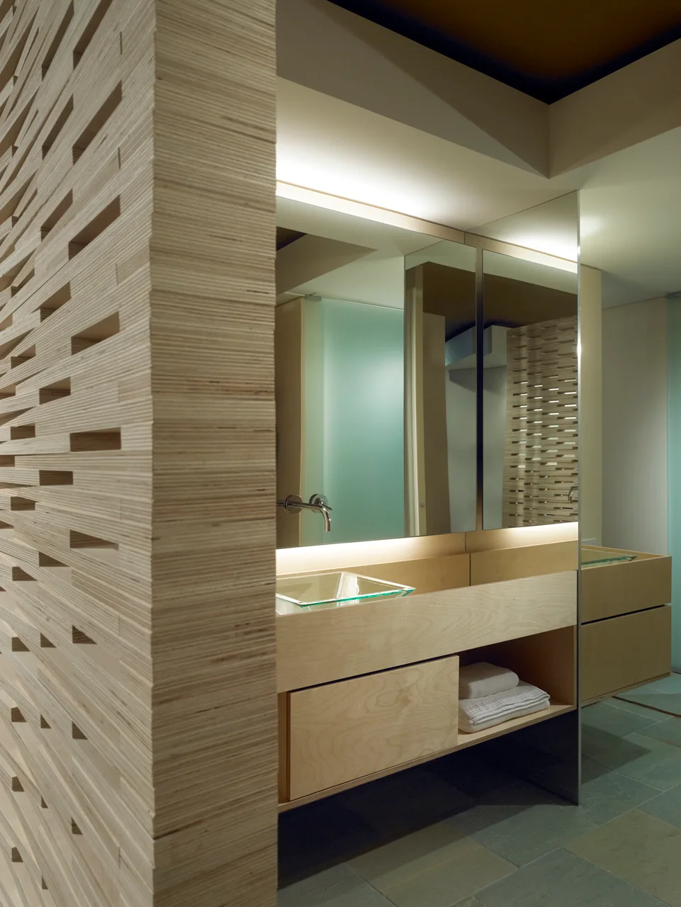 A modern bathroom featuring a slatted plywood wall partition, a light wood floating vanity with a glass basin sink, and a large mirror.