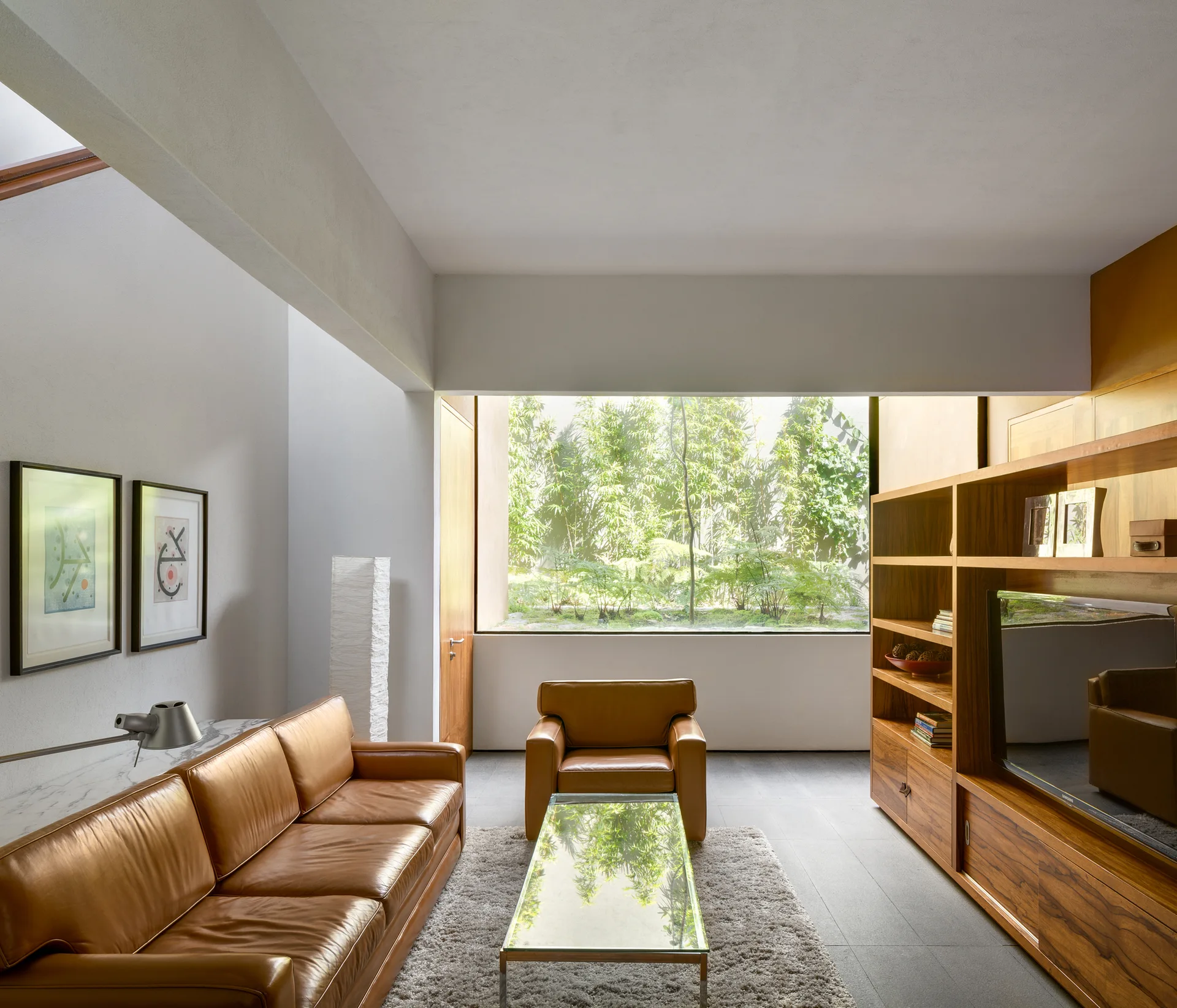 A modern living room features a tan leather sofa, matching armchair, a glass coffee table, and a large window showcasing a lush green garden.