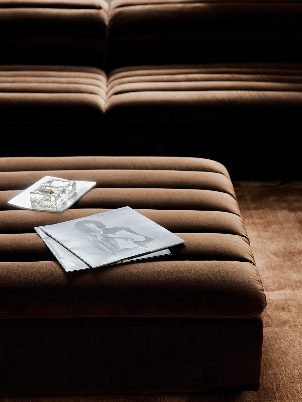 A brown channeled velvet ottoman holding a glass tray and a book sits in front of a matching sofa blurred in the background.
