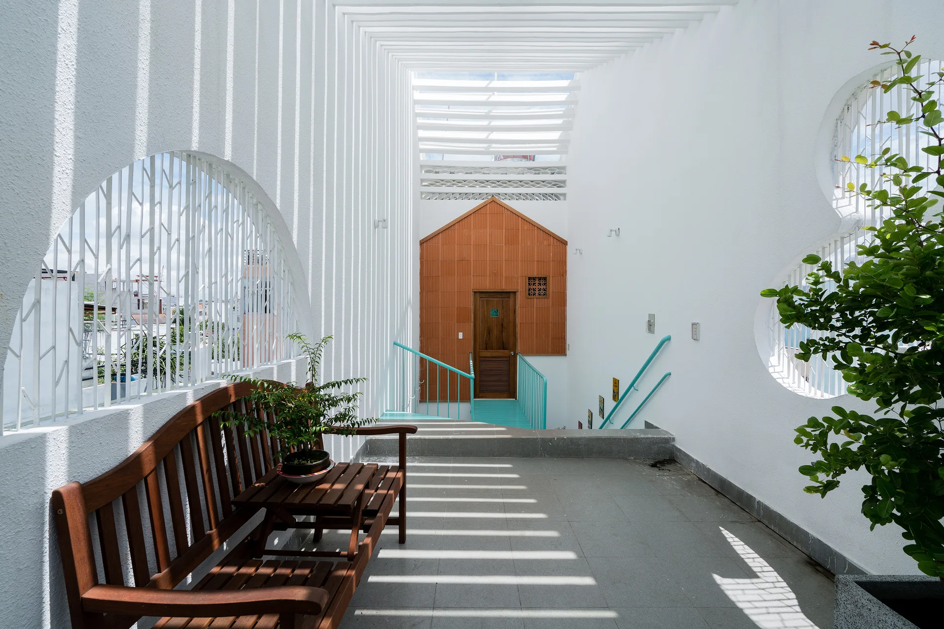 A sunlit white stucco terrace features a wooden bench, decorative wall cutouts, a teal metal handrail, and dramatic shadows cast by a slatted pergola ceiling.