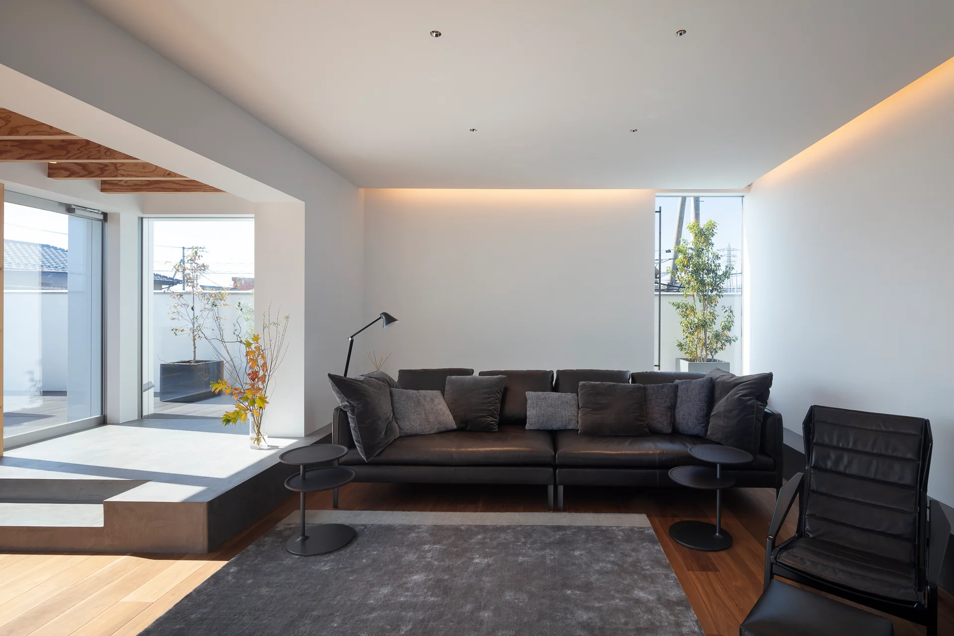 A minimalist living room features a dark leather sofa on hardwood floors next to a raised concrete platform and large glass windows.