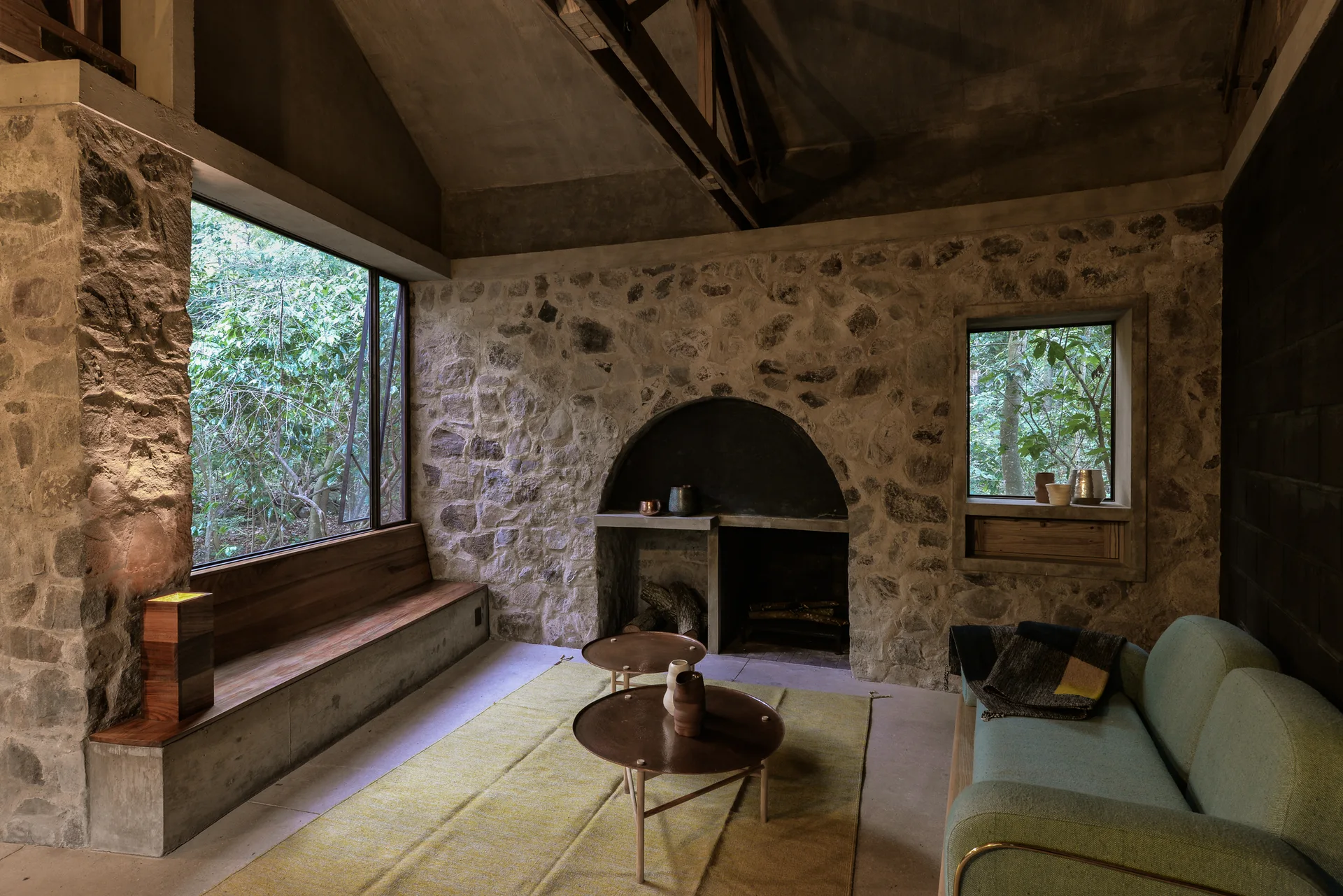 A rustic living room features textured stone walls, an arched fireplace, a built-in wooden bench, and a light green sofa near a large window.