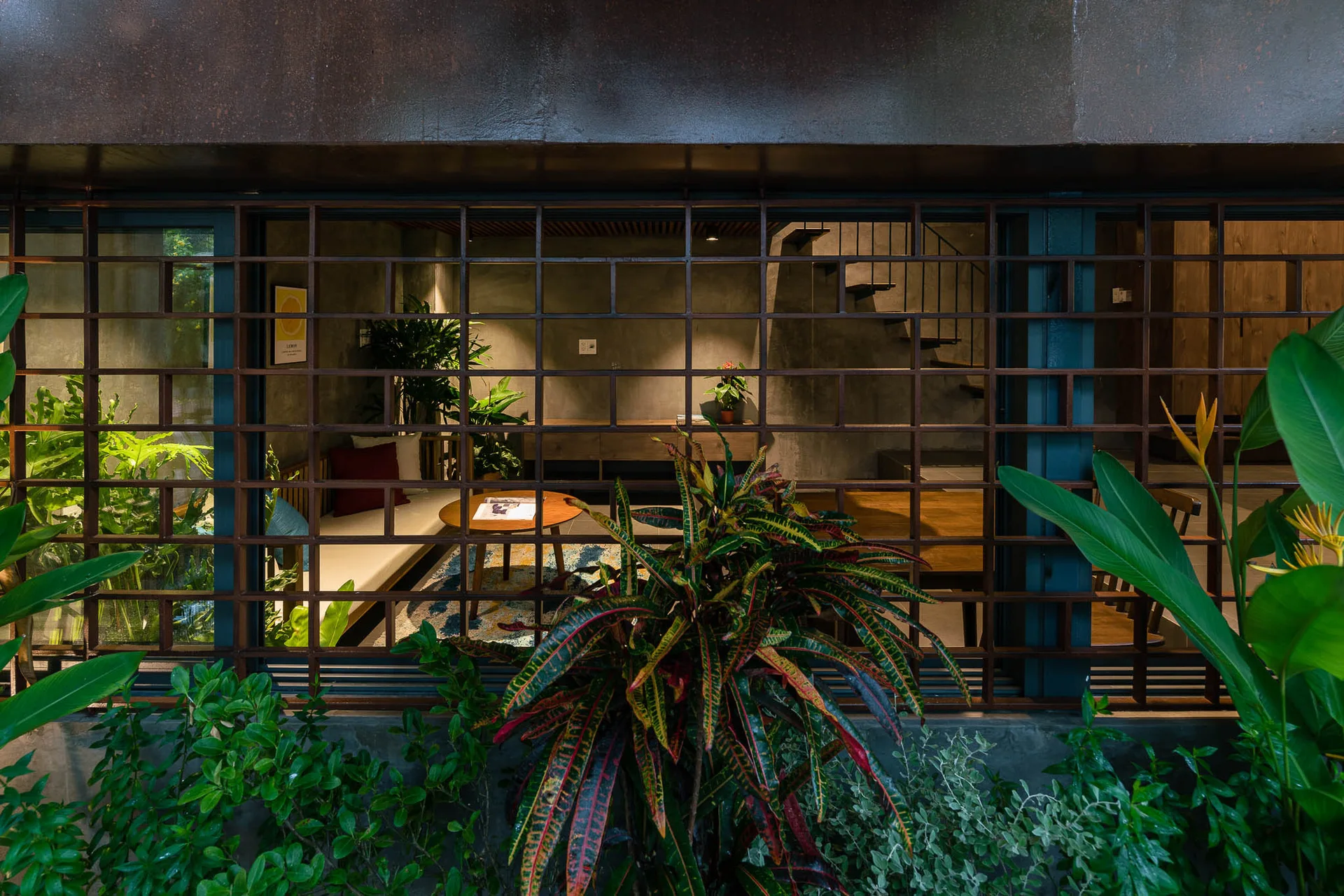 A view through a dark metal window grid into a minimalist living room with concrete walls, a low wooden sofa, and indoor plants.
